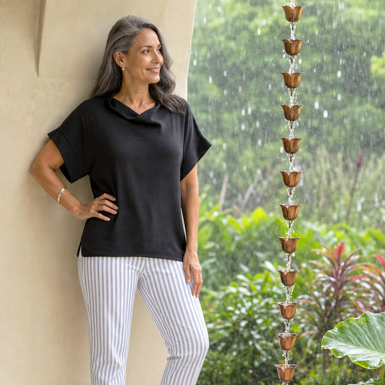 Woman leaning against the wall with rain behind her wearing a black top and striped pants.
