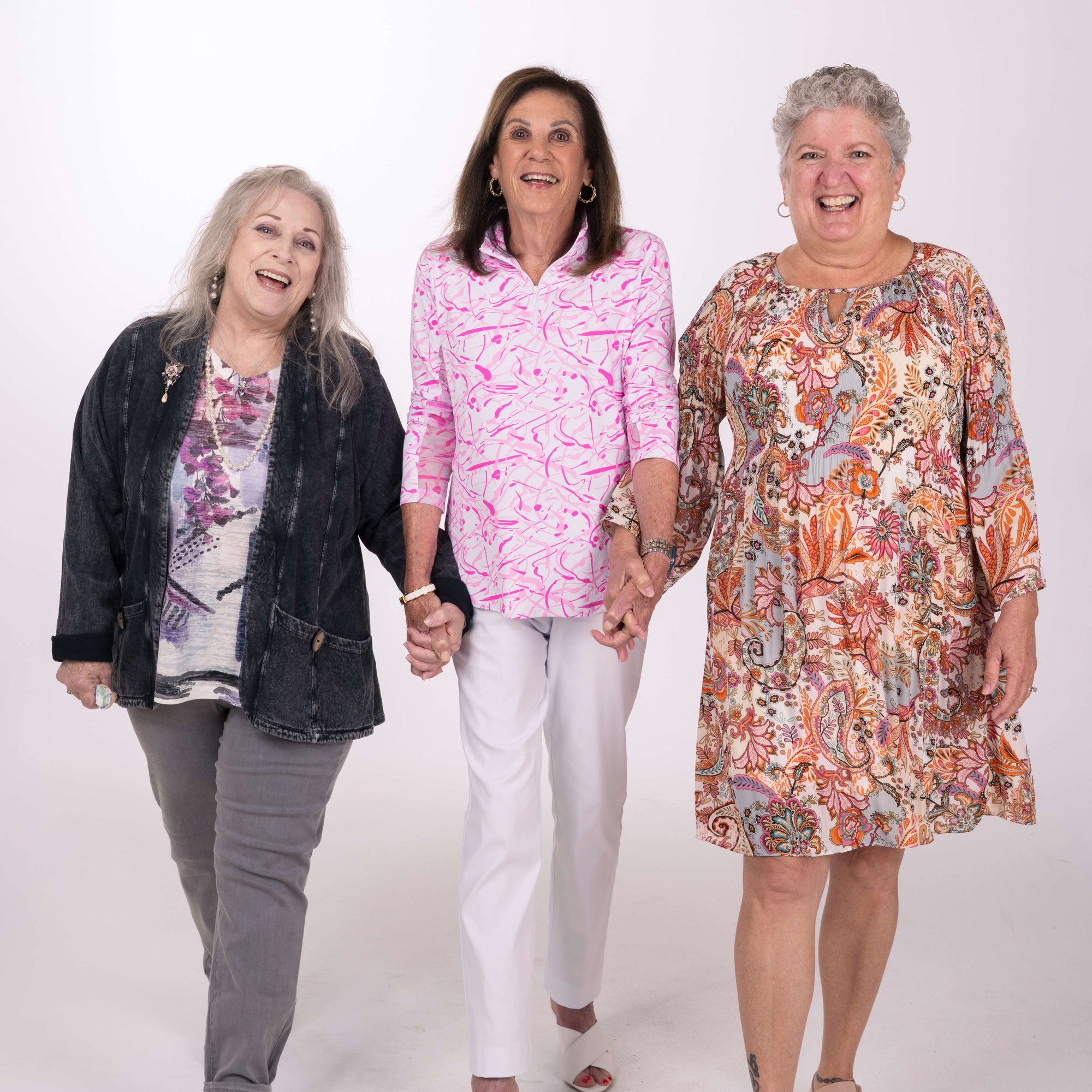 Three smiling women hold hands as they walk forward in front of a white studio background.