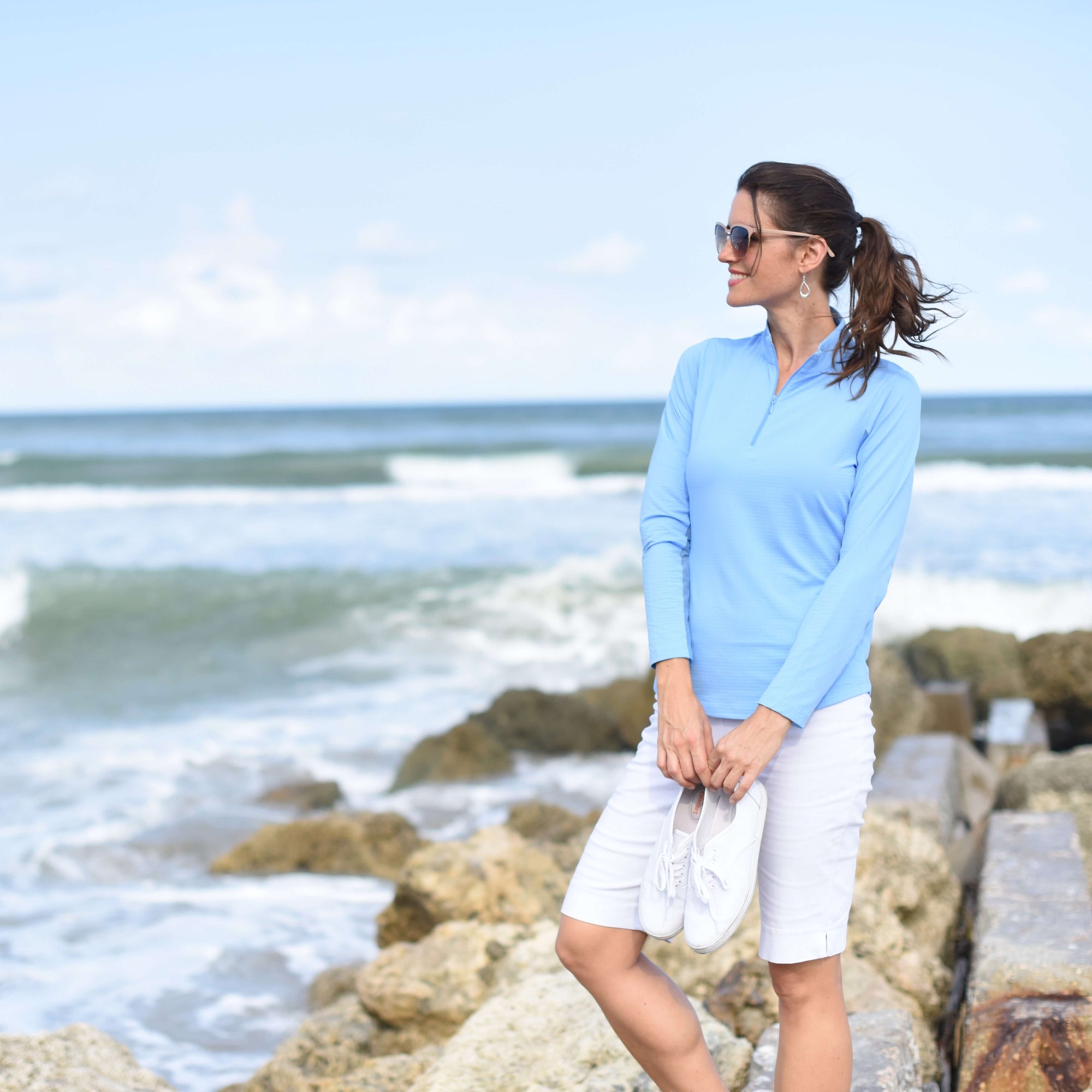 A woman stands on a rocky shoreline wearing a light blue long-sleeve polo shirt and white shorts, holding white sneakers, with the ocean and sky in the background.