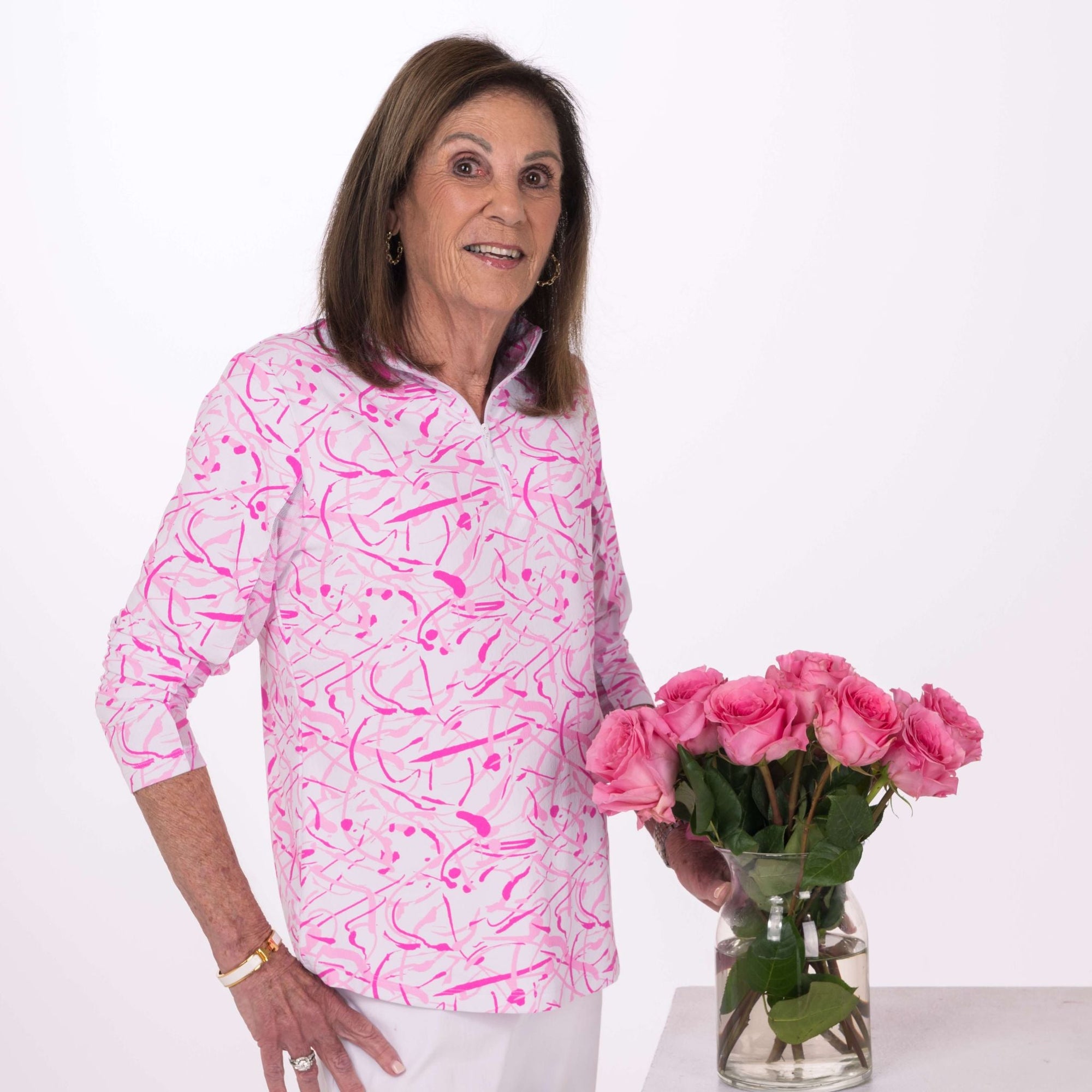 A woman wearing a white and pink sun shirt stands next to pink roses against a light colored studio background