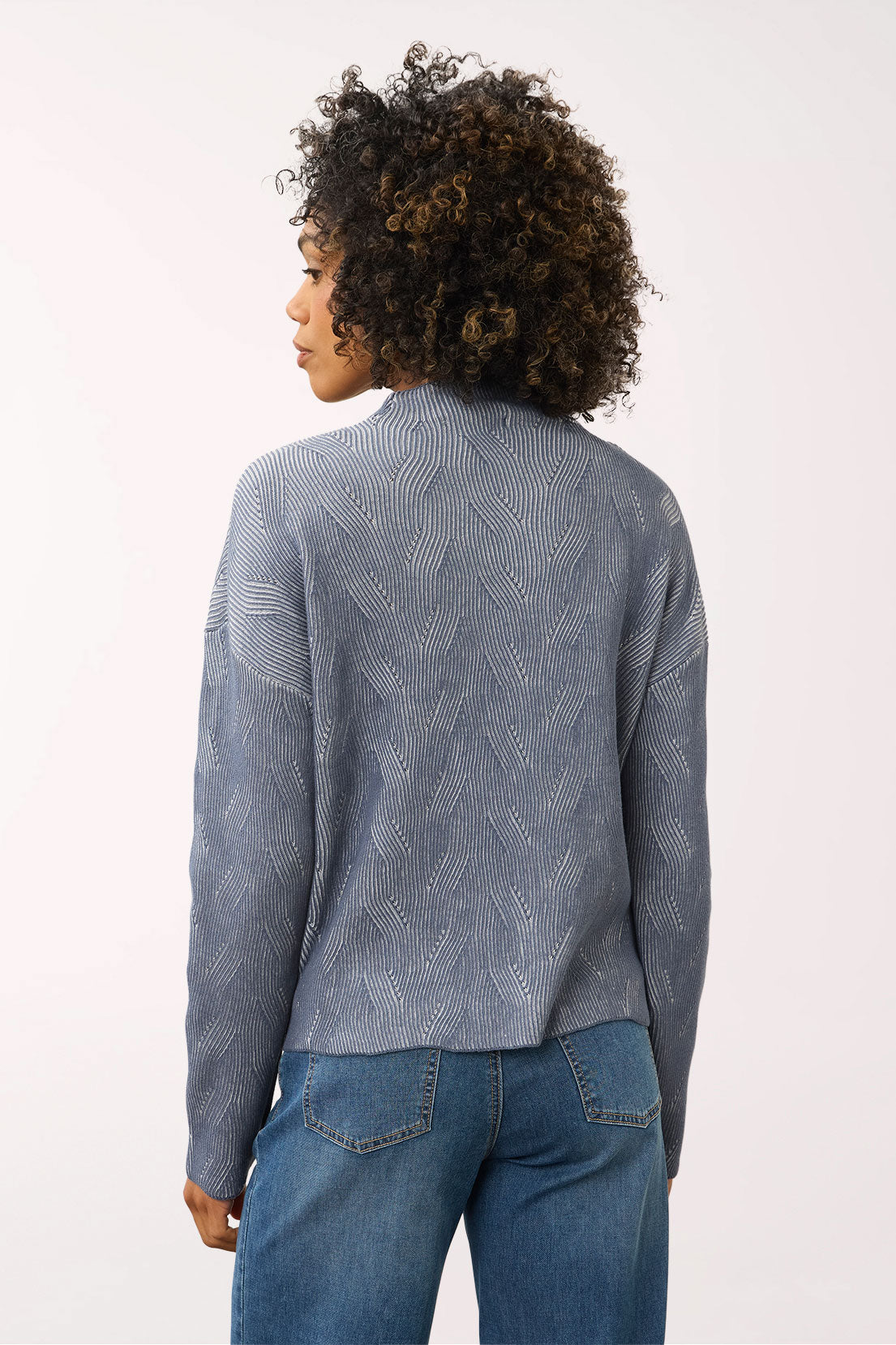 A woman shown from the back wearing a textured, patterned gray-blue knit sweater and blue jeans against a plain white studio background.