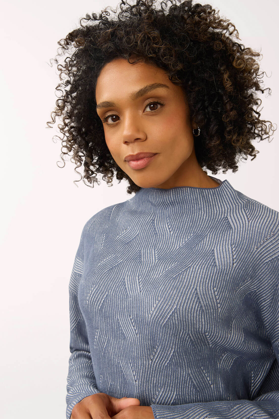 A woman with curly dark hair wearing a textured blue-gray long-sleeve top, photographed against a plain light background.