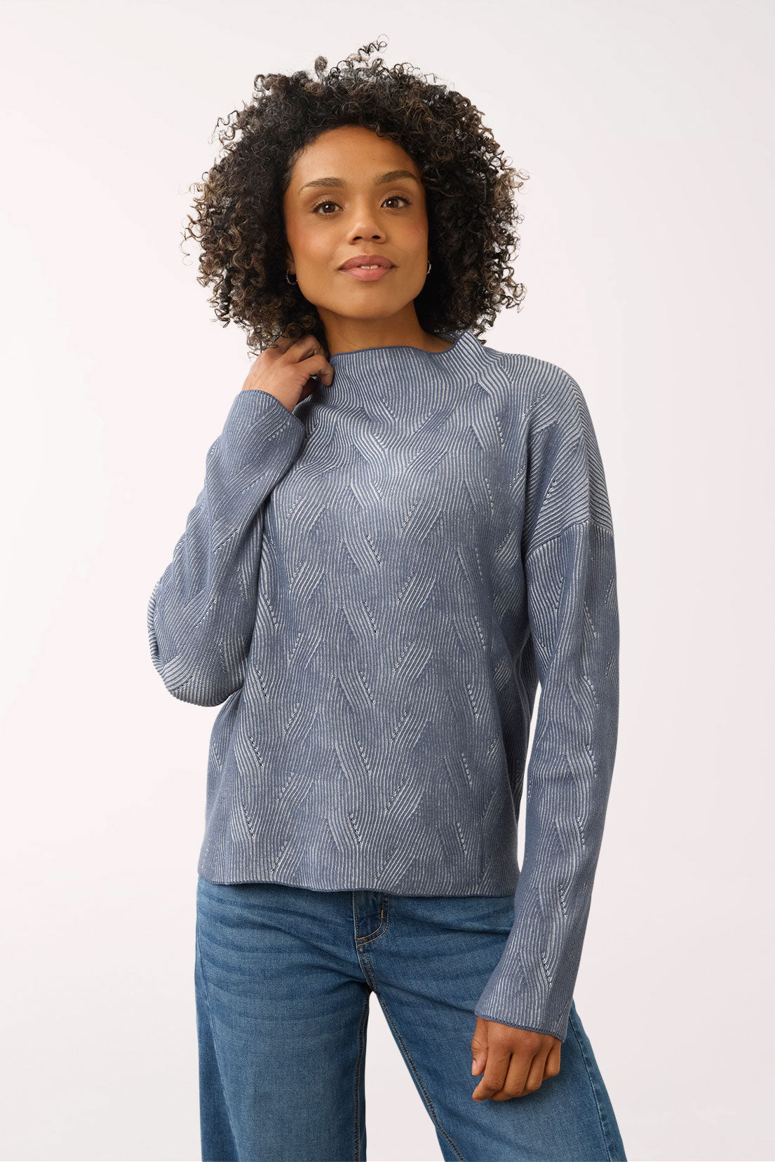 A woman wearing a textured, long-sleeve knit sweater in a dusty slate blue-gray color, paired with blue jeans, against a plain white studio background.