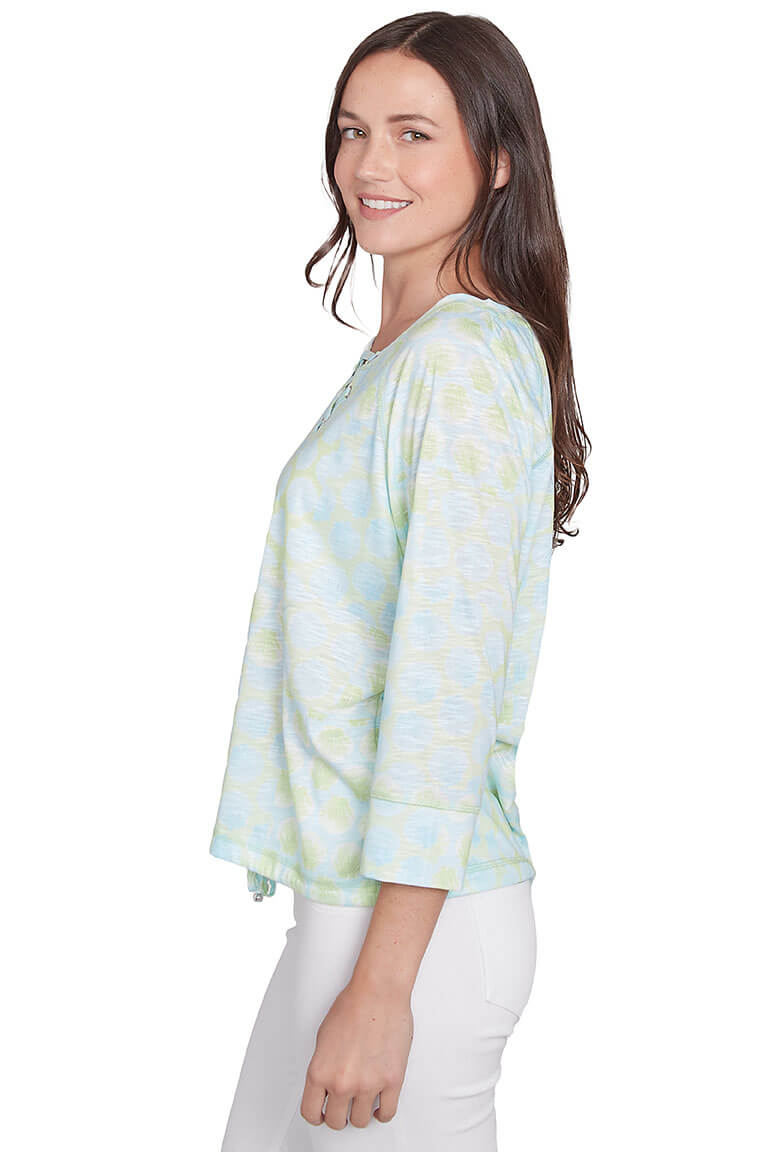 A side shot of a model wearing a petite-sized, floral print, 3/4 raglan sleeve top with a split v-neckline and lace-up detail.