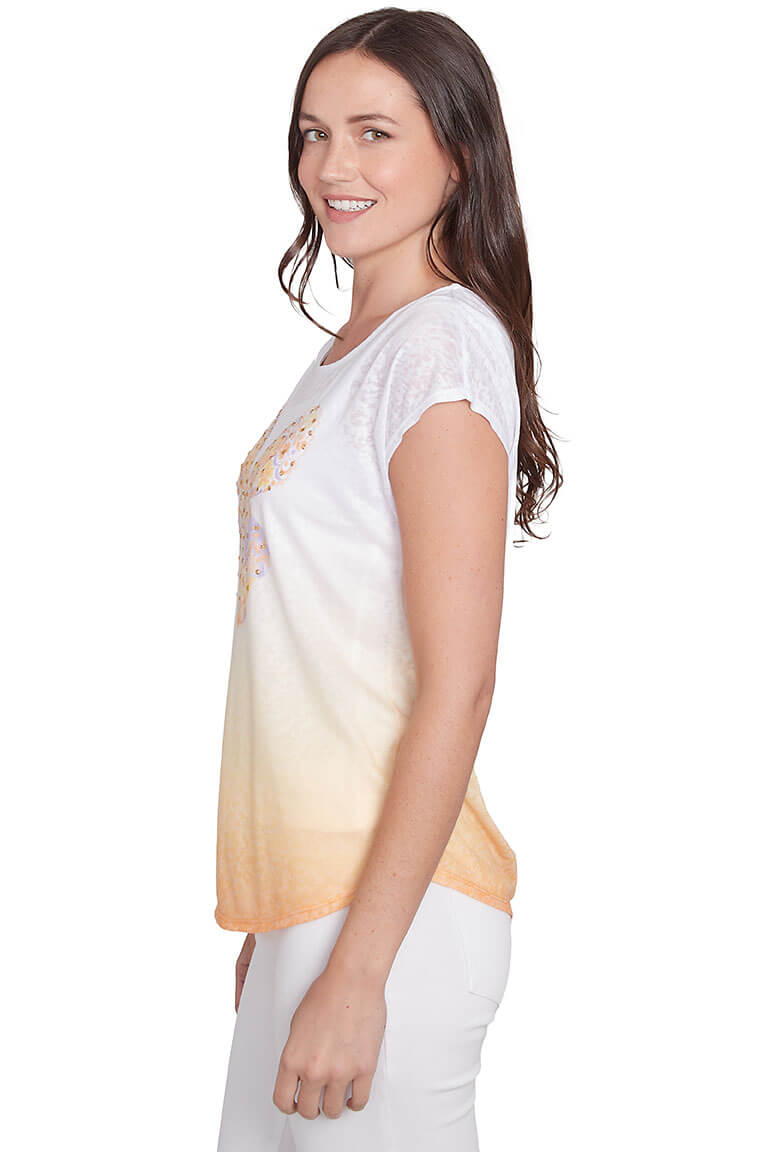 A side shot of a model wearing a white scoop neck tee with short dolman sleeves and a curved hemline. The tee features a peach and orange gradient color with a jeweled butterfly embellishment on the chest.
