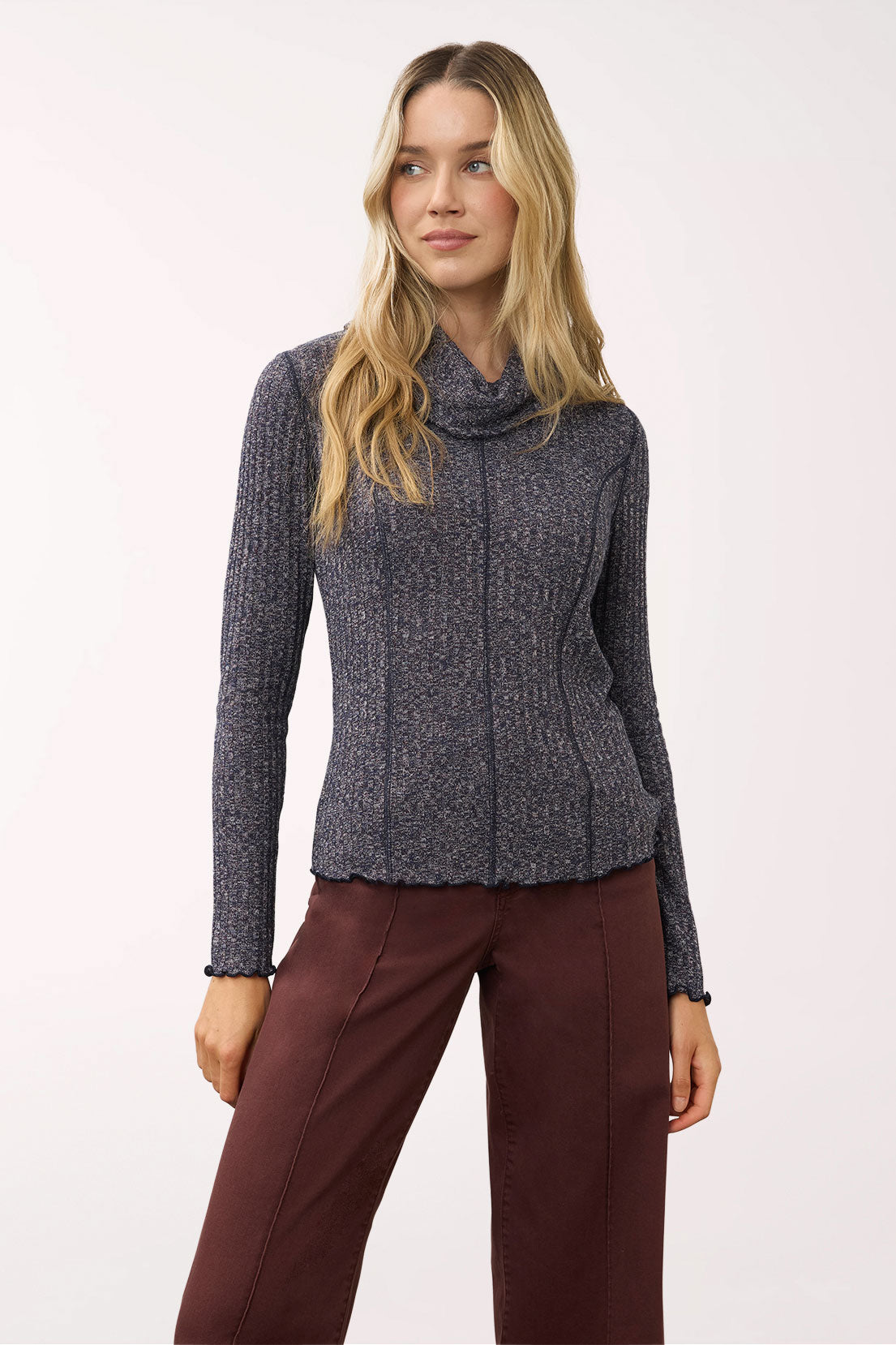 A woman modeling a dark gray ribbed knit cowl-neck sweater paired with dark brown pants, photographed against a plain light background.