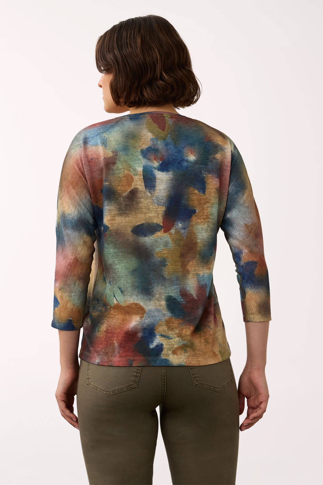 A woman viewed from the back wearing a three-quarter sleeve top with an abstract watercolor-style print in blue, teal, brown, and rust tones, paired with olive green pants against a plain white studio background.