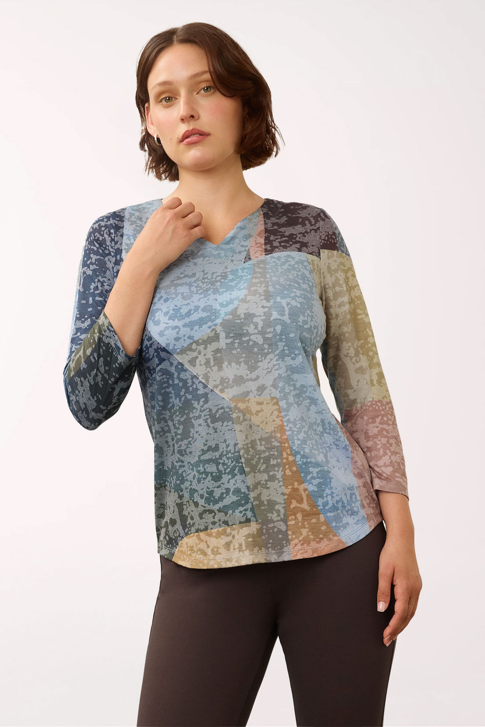 A woman modeling a long-sleeve V-neck top with a multicolor abstract patchwork print in shades of blue, beige, brown, and gray, paired with dark brown leggings, against a plain white studio background.