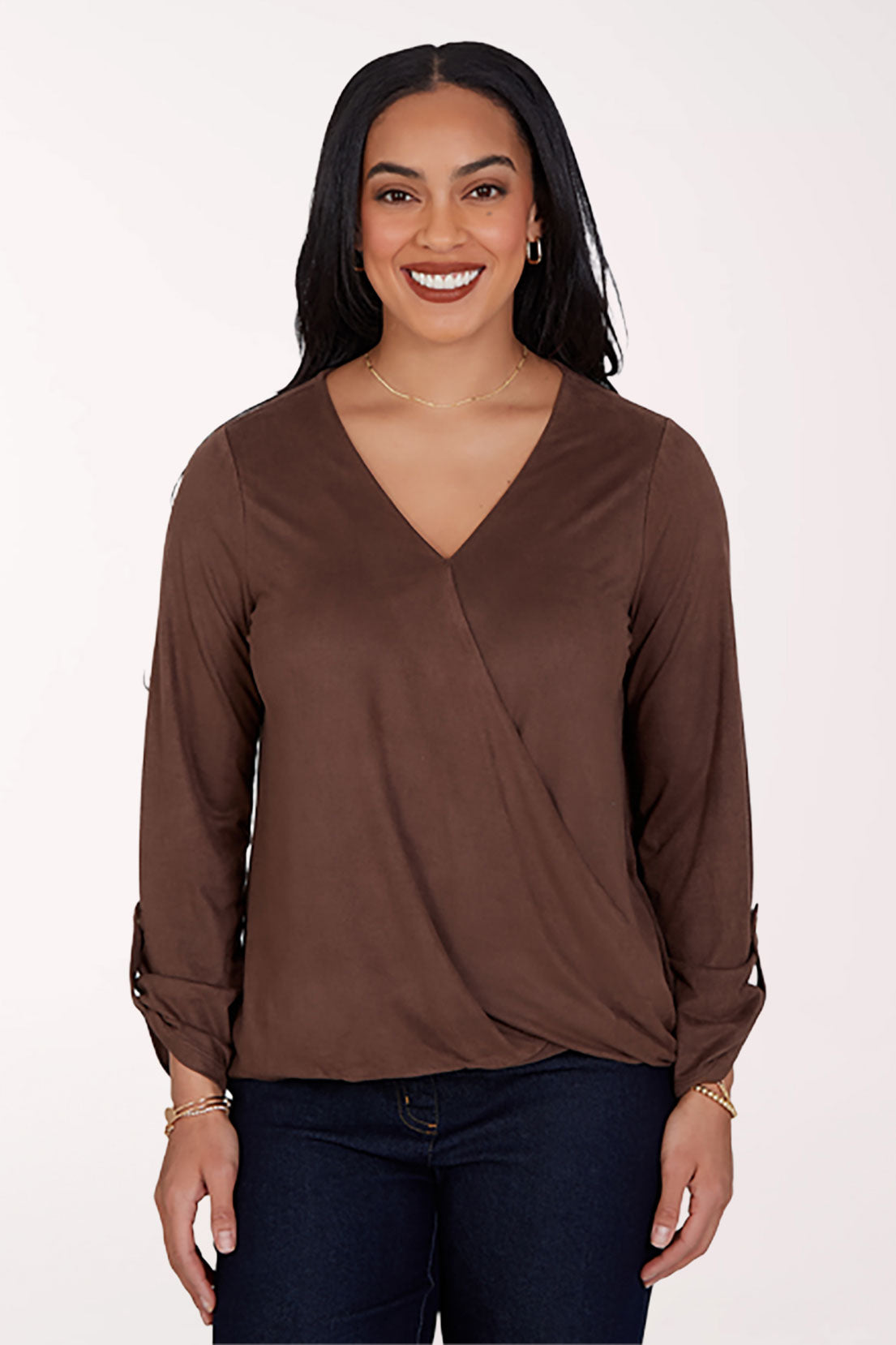 A woman modeling a brown long-sleeve wrap-style blouse with a V-neckline, paired with dark blue jeans, against a plain light background.