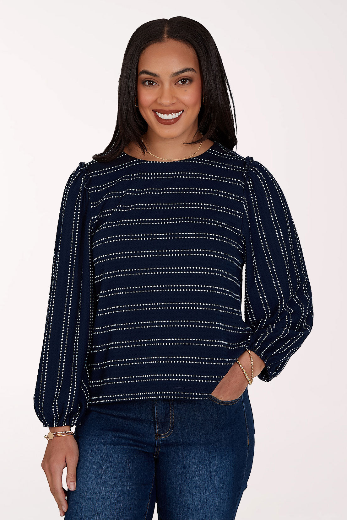 A woman modeling a navy blue long-sleeve top with thin white dotted horizontal stripes, paired with dark blue jeans, against a plain white studio background.
