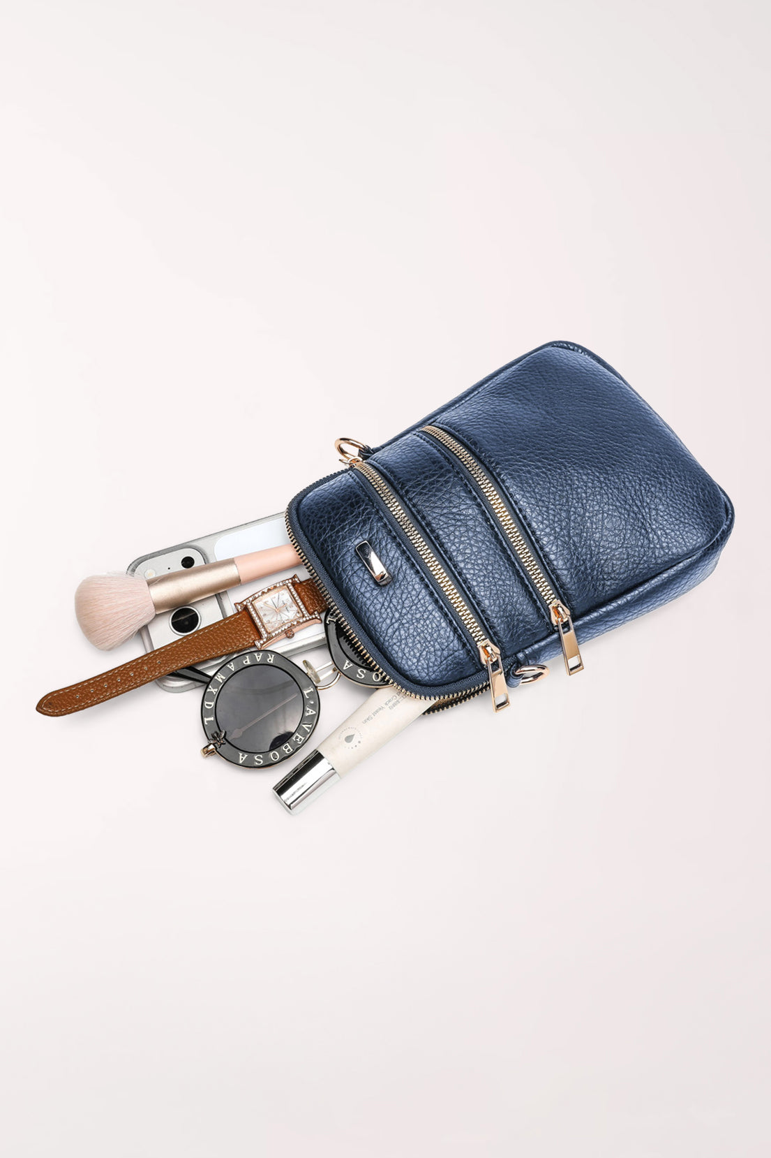 The navy 22 Tote Three-Pocket Crossbody Bag lying on its side with a phone, makeup brush, compact mirror, lipstick and watch spilling out to show what fits inside.