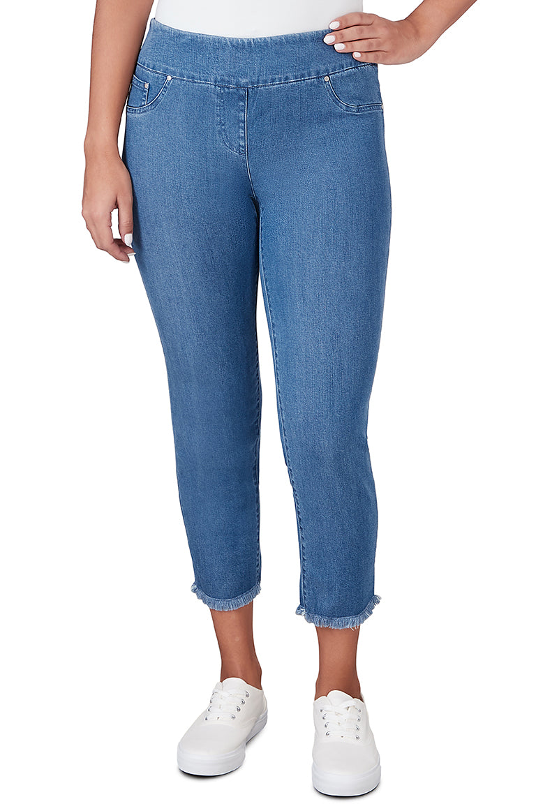 A front shot of a model wearing a pair of medium blue fringed denim ankle pants with an elastic waistband, faux front pockets, and frayed hem. The pants are pull-on style and the model is wearing white sneakers.