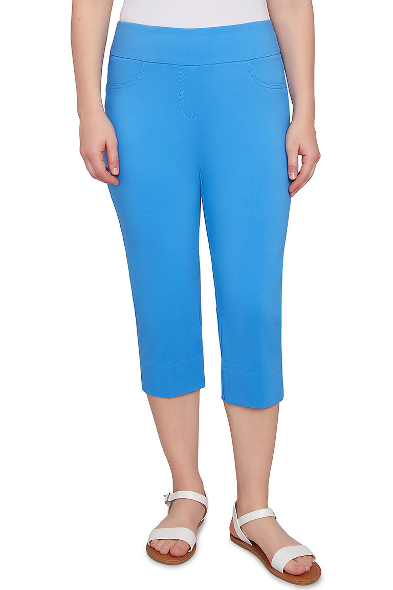 A front shot of a model pair of blue pull-on capri pants with an elasticized waistband, faux front pockets, back patch pockets, and lattice detail on the hemline.