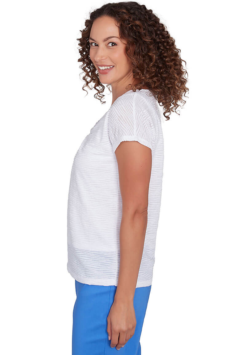 A side shot of a model wearing a white cap sleeve knit top with a V-neckline, ringed styling, and rhinestone studded embellishment. The top has a straight hemline and wave-textured fabric.