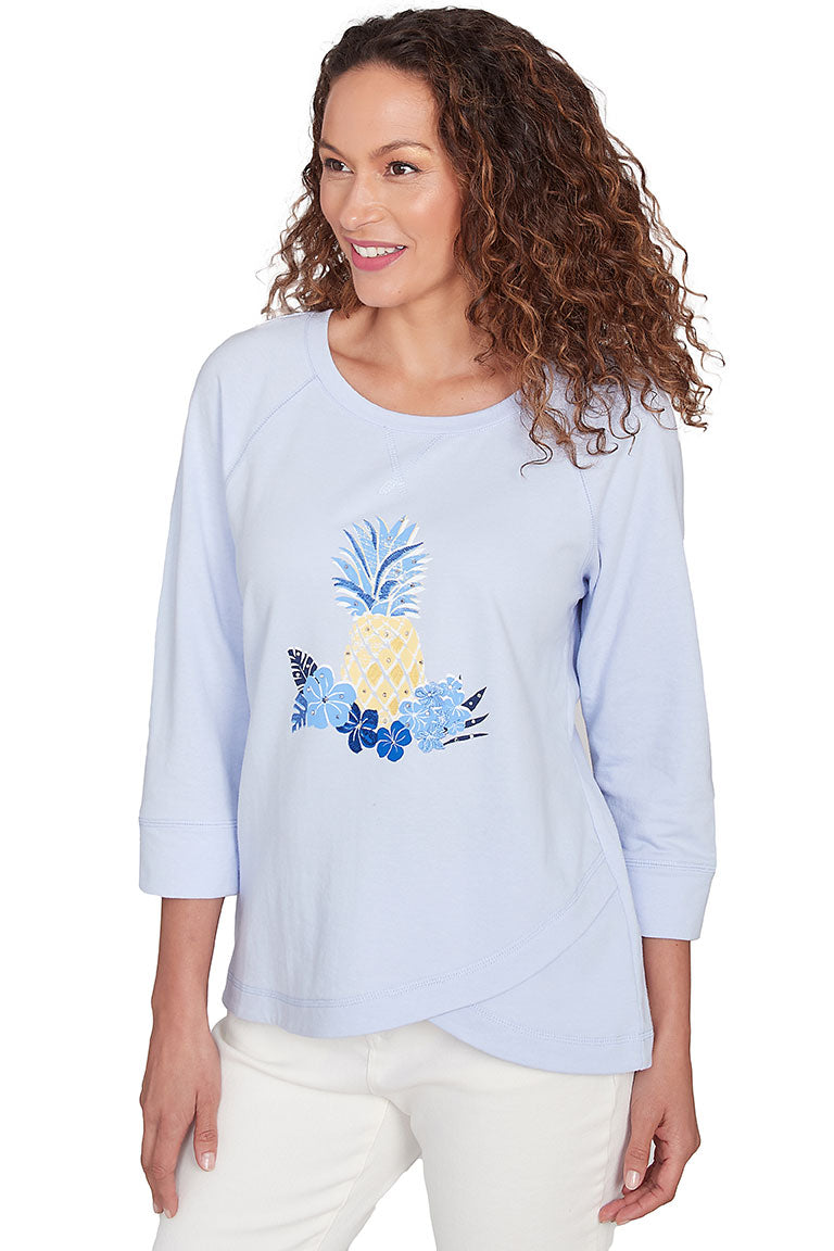A front shot of a model wearing a light blue, scoop neck knit top with 3/4 raglan sleeves, an embroidered design with rhinestone embellishment, and an asymmetrical tulip hemline.