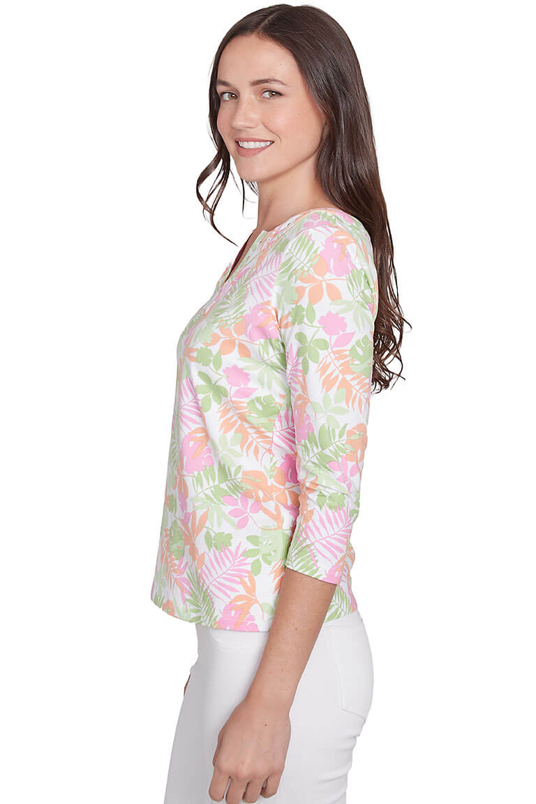 A side shot of a model wearing a floral print pink and green 3/4 sleeve top with a notched v-neckline and studded embellishment, paired with white pants.