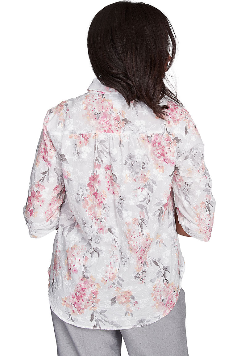 A back shot of a woman wearing a white, pink, and green floral button-front jacquard blouse with a foldover collar, pleated shoulders, and 3/4 sleeves with slits on the cuffs.