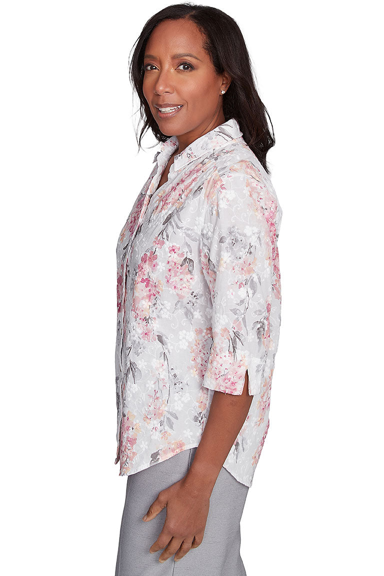 A side shot of a woman wearing a white, pink, and green floral button-front jacquard blouse with a foldover collar, pleated shoulders, and 3/4 sleeves with slits on the cuffs.