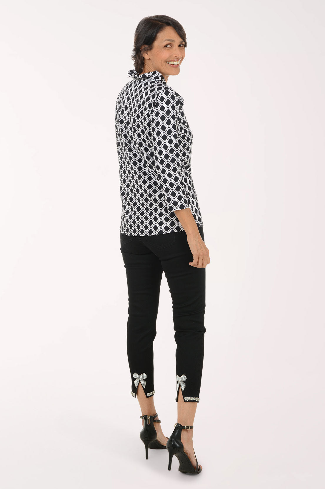 A woman modeling a black and white geometric patterned long-sleeve blouse with black cropped pants featuring bow details at the cuffs, and black high heels, photographed against a plain white studio background.