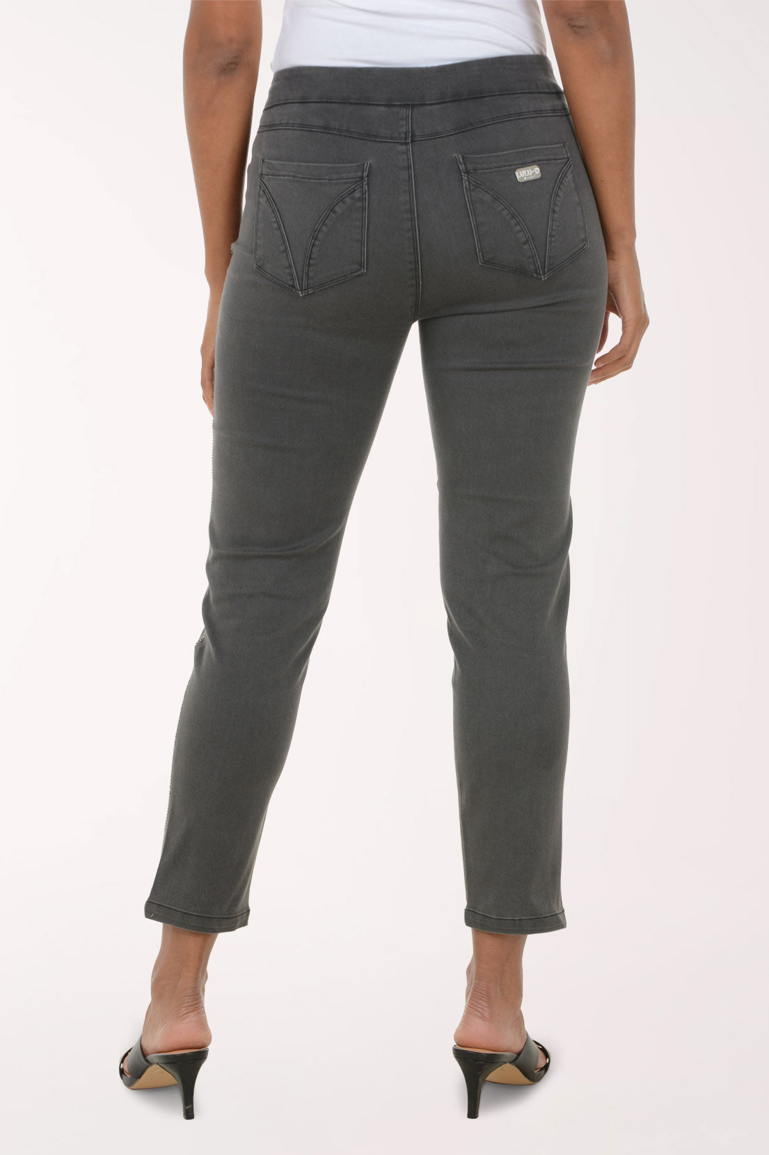 A model wearing a pair of dark gray slim-fit cropped jeans with a straight leg, paired with a white top and black heeled sandals, against a plain white studio background.
