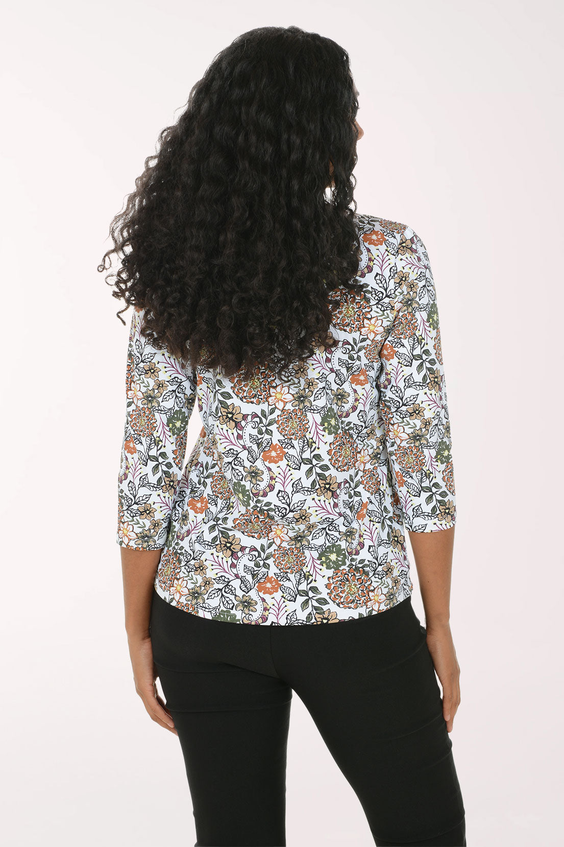 A woman with long curly black hair wearing a white three-quarter sleeve top with a colorful floral and paisley pattern, paired with black pants, standing against a plain white background.