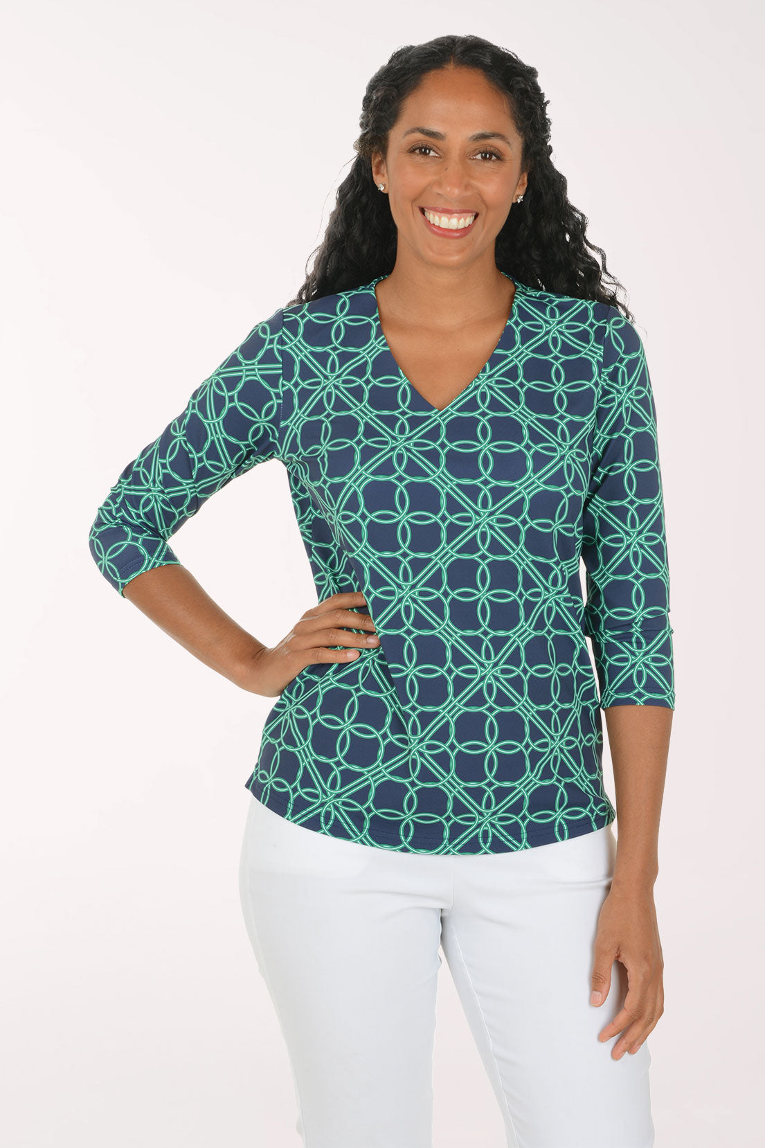 A woman modeling a three-quarter sleeve V-neck top with a teal and navy geometric interlocking circle pattern, paired with white pants, against a plain white studio background.