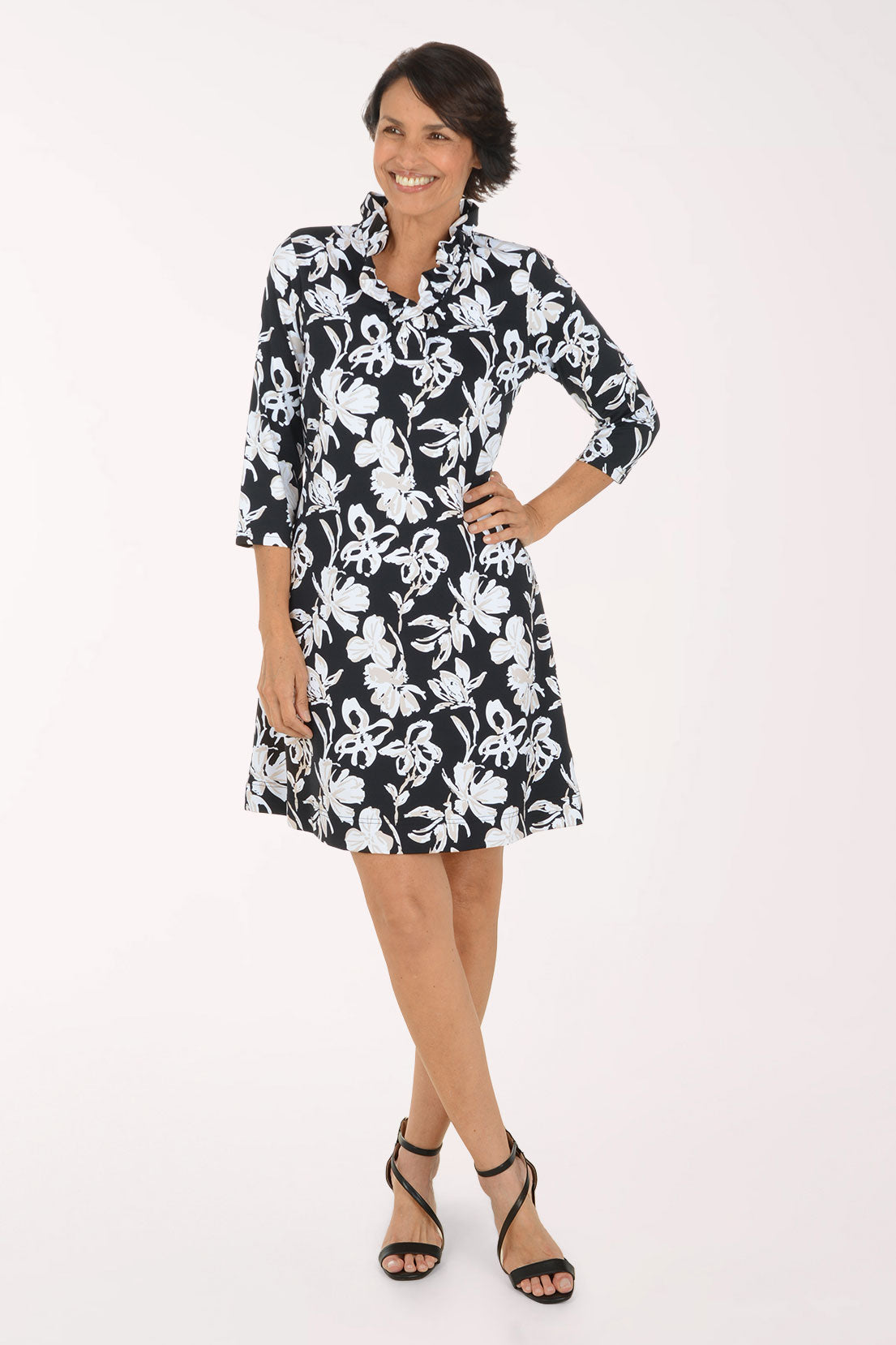 A woman modeling a black and white floral print shirt dress with three-quarter sleeves and a collared neckline, paired with black strappy high-heeled sandals against a plain white studio background.
