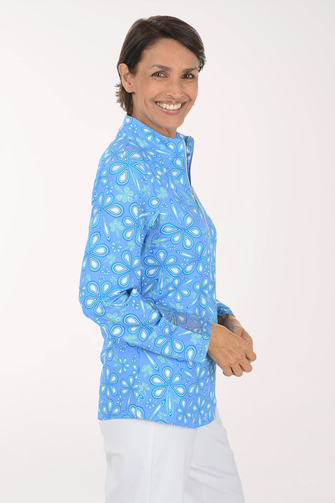 A smiling woman with short brown hair stands against a light off-white studio background, wearing a light blue long-sleeve quarter-zip pullover with a white paisley pattern and white pants.