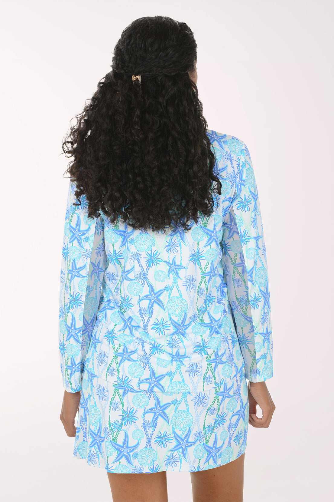 A smiling woman wearing a matching two-piece lounge set with a light blue and white starfish and palm tree print, featuring a long-sleeve quarter-zip top and a matching skirt.
