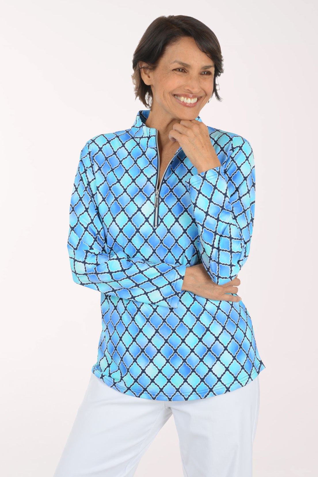 A woman modeling a blue and light blue diamond-patterned quarter-zip pullover top with white pants, set against a plain white studio background.