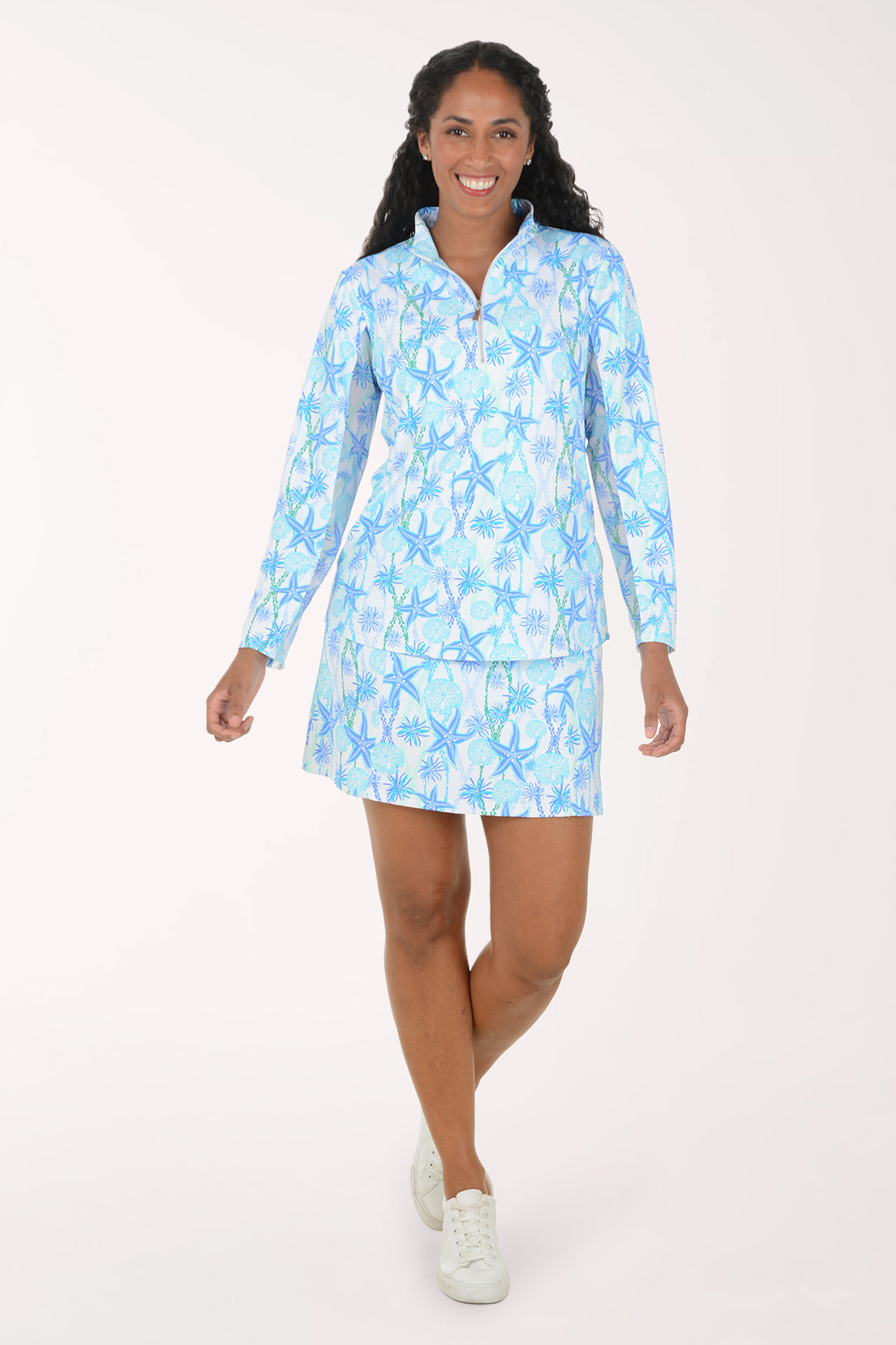 A woman modeling a light blue and white patterned long-sleeve mini dress with a half-zip collar, set against a plain light background.