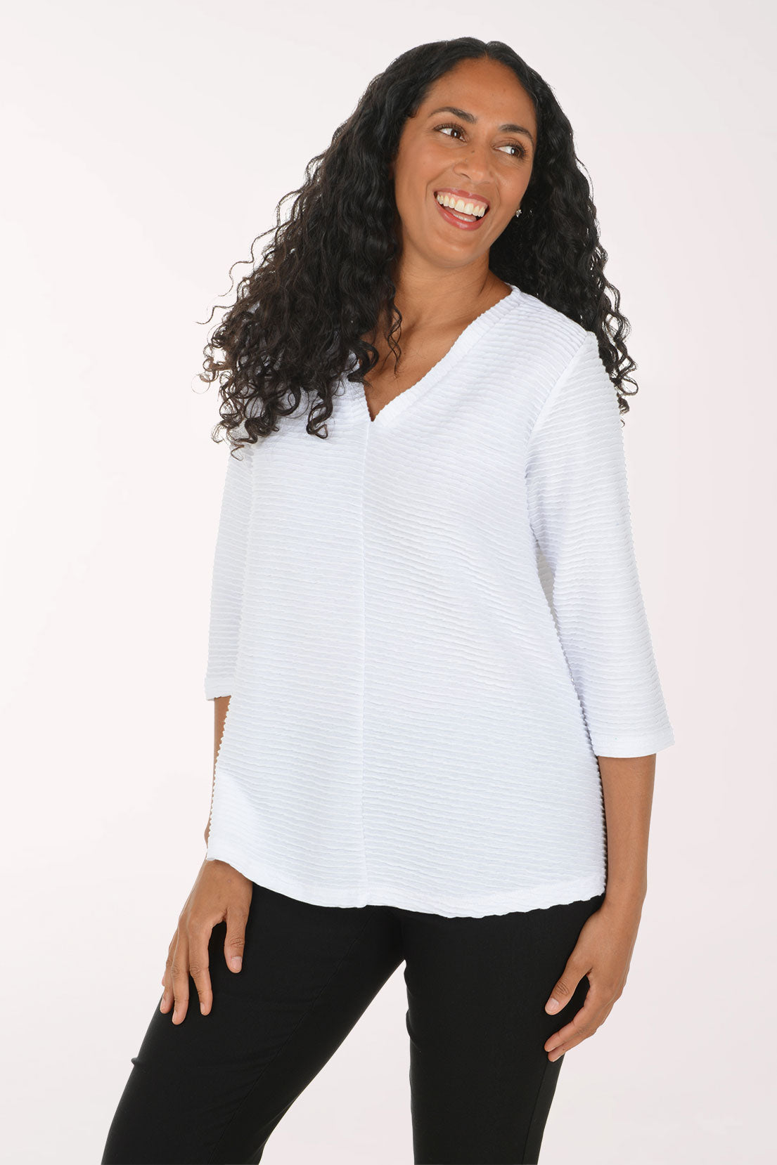 A woman with long curly black hair wearing a white textured three-quarter sleeve top and black pants, posing against a plain white studio background.