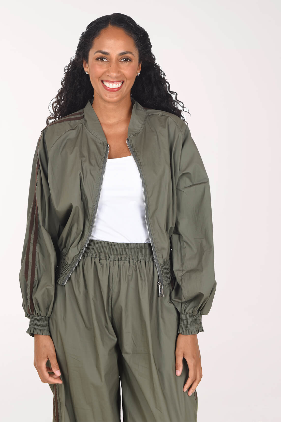 A smiling woman wearing an olive green lightweight zip-front jacket with dark brown stripes on the sleeves, matching olive green jogger pants, and a white tank top, photographed against a plain light background.