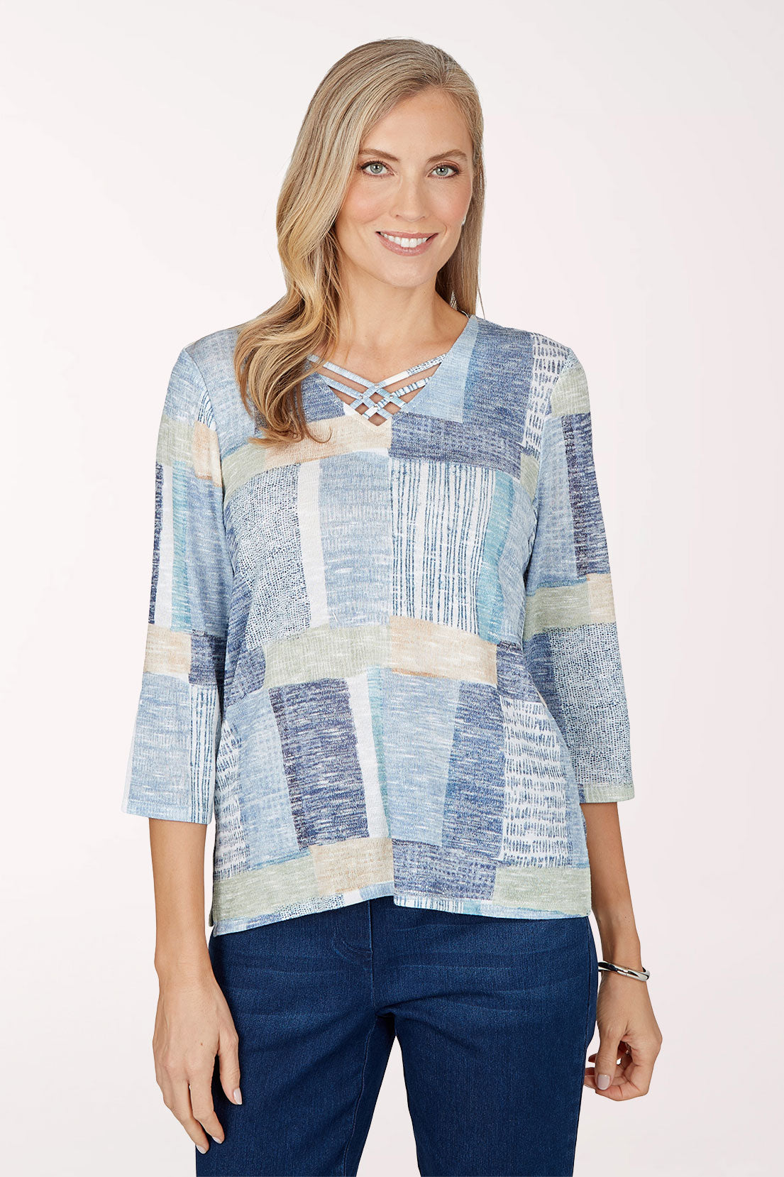 Front view of a woman in a knit top with a patchwork print of navy, sky blue, white and cream textured blocks, a criss-cross V-neckline and 3/4 sleeves, worn with dark jeans.