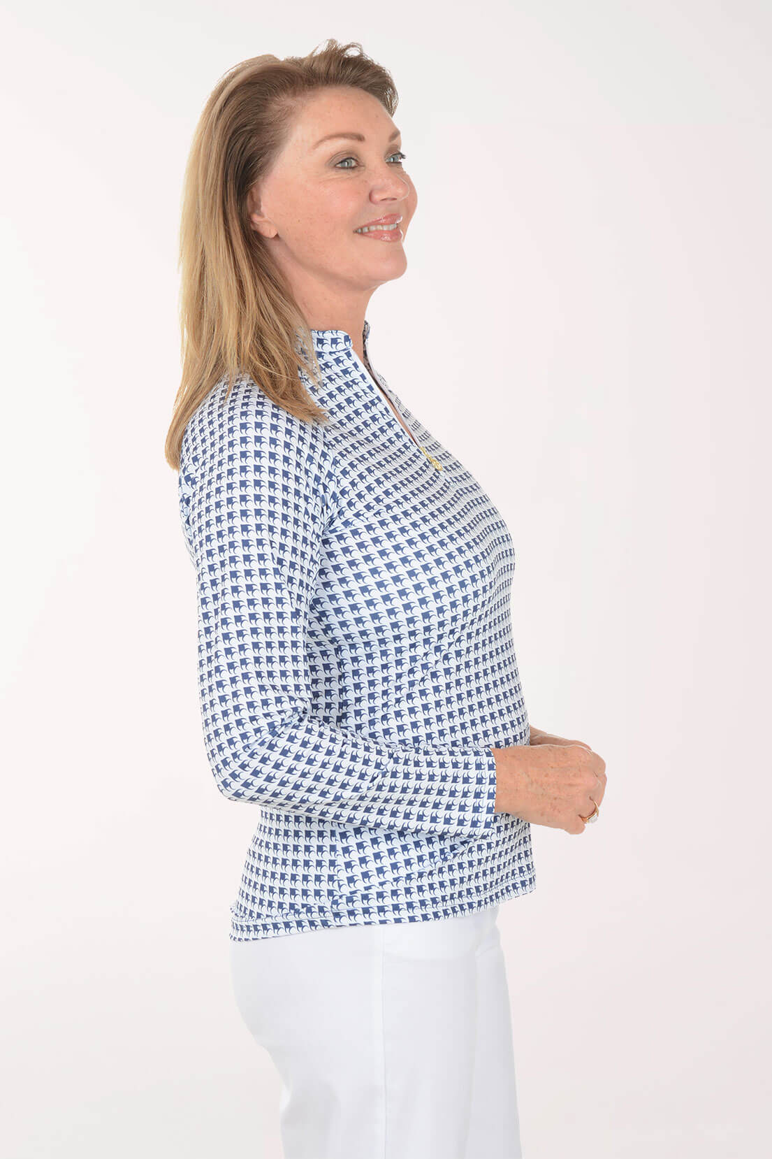 A woman modeling a long-sleeve mock neck top with a blue and white geometric pattern, paired with white pants, against a plain white studio background.