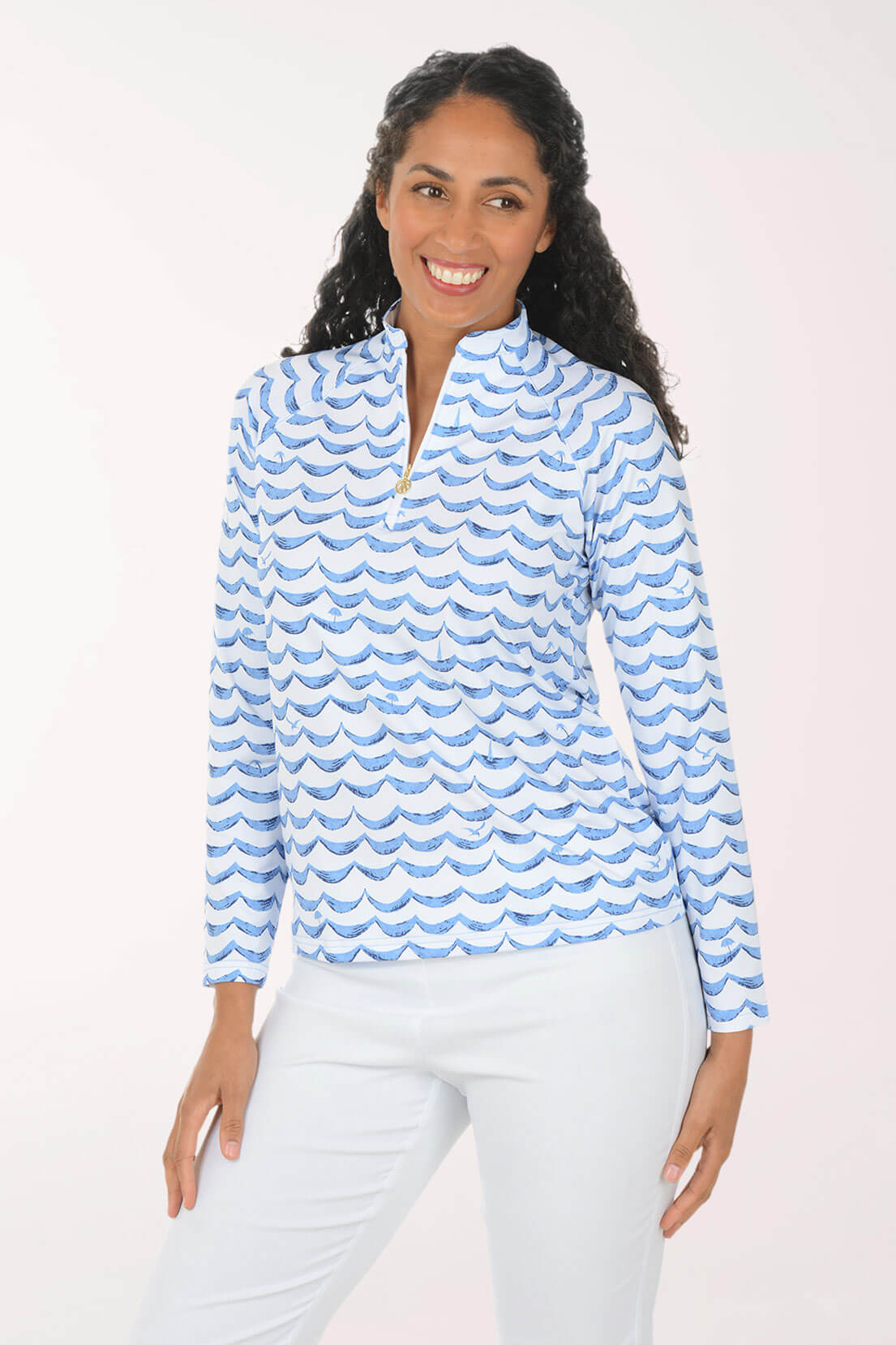 A woman modeling a light blue and white wavy patterned long-sleeve quarter-zip pullover top, paired with white pants, against a solid pale pink studio background.