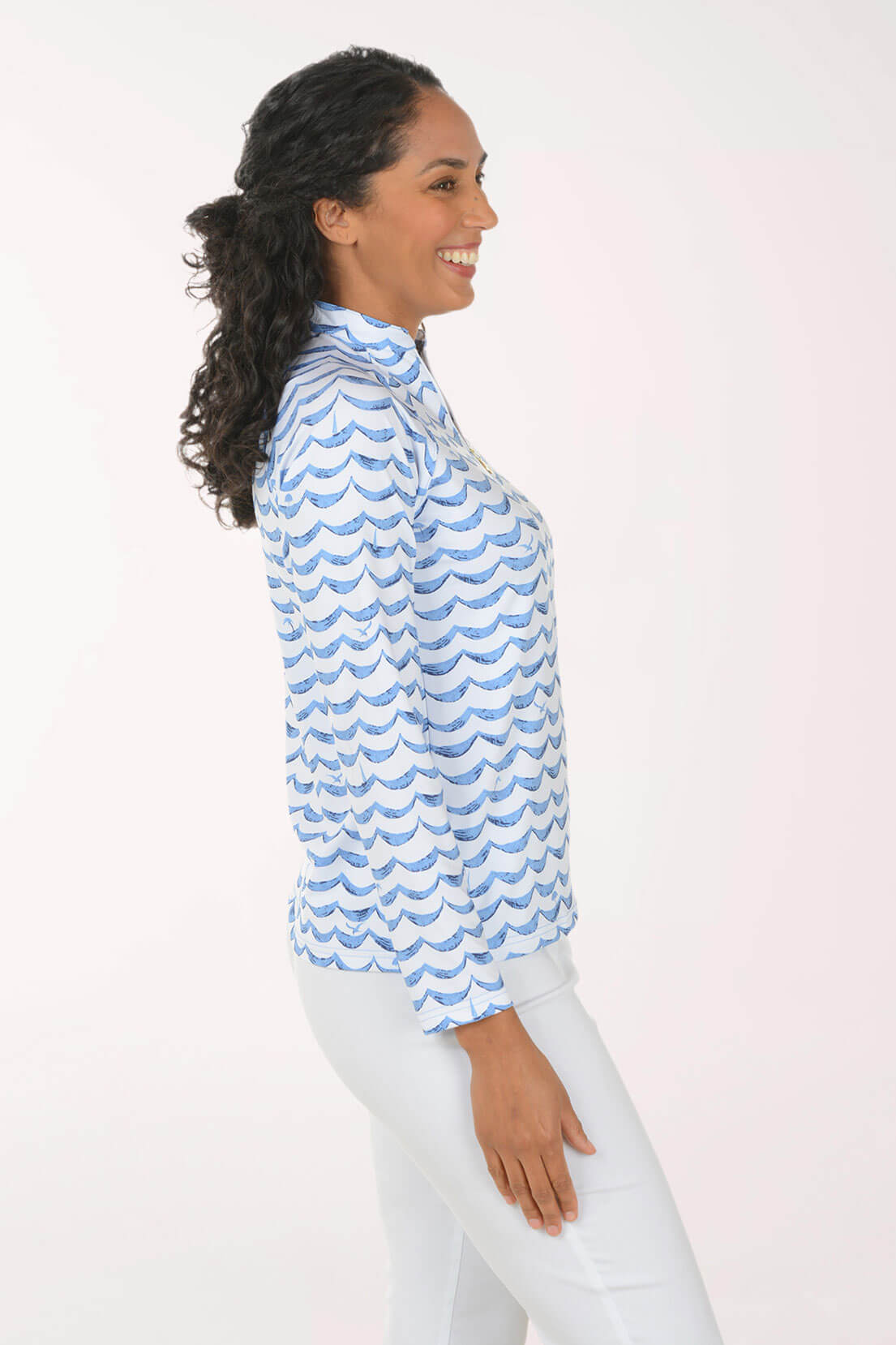 A woman shown in side profile wearing a light blue and white patterned long-sleeve blouse with white pants, standing against a plain light background.