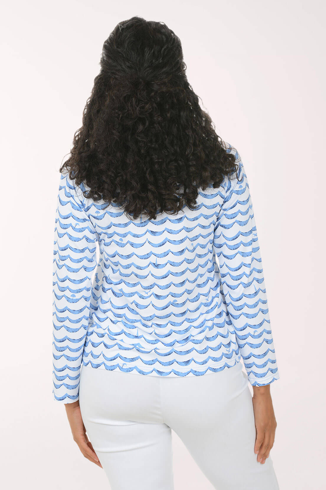 A woman viewed from behind wearing a light blue and white patterned long-sleeve top and white pants against a plain light background.