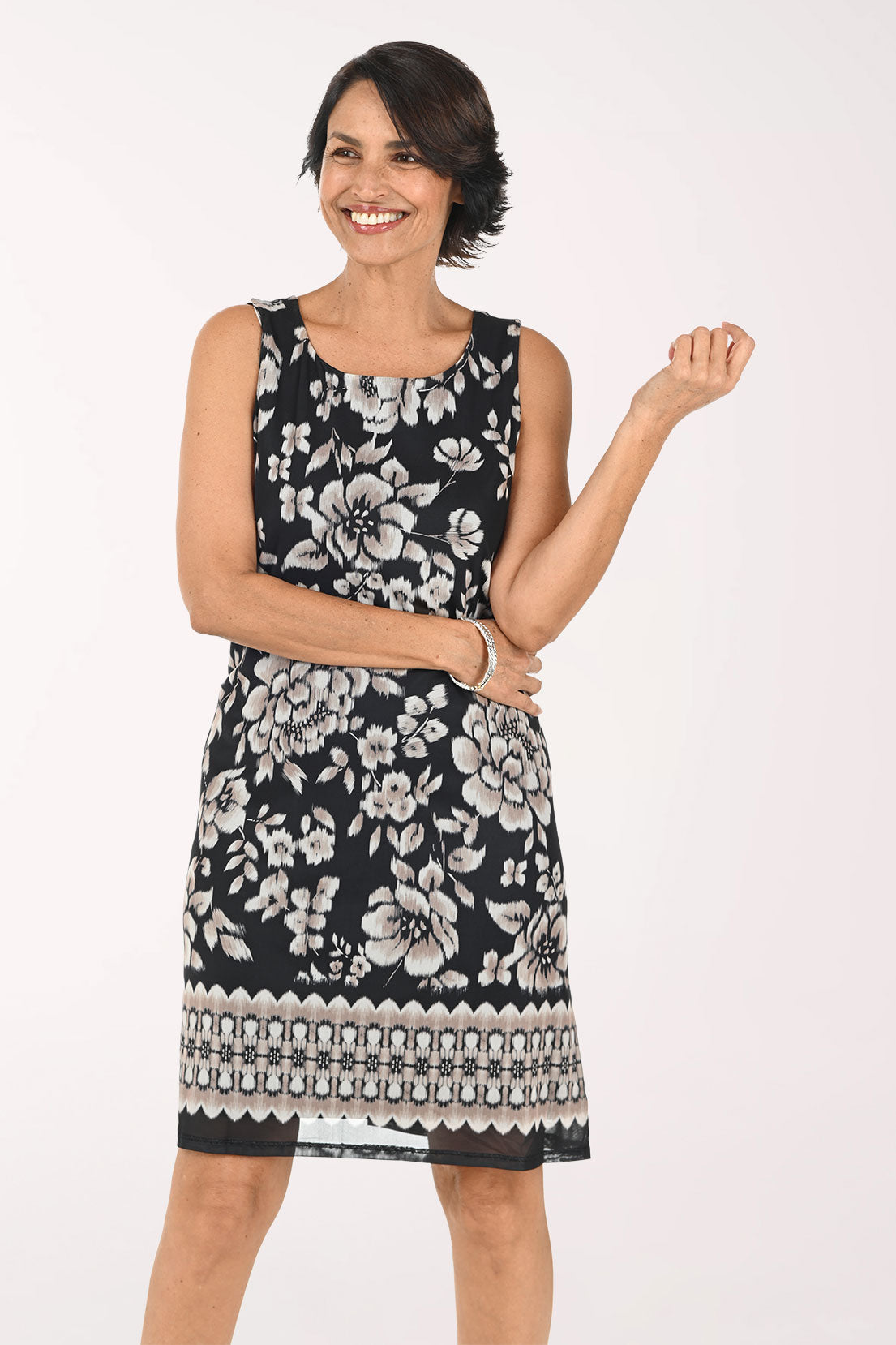 A woman modeling a sleeveless black and white floral print dress with a geometric patterned hem, paired with black strappy heels, against a plain white studio background.