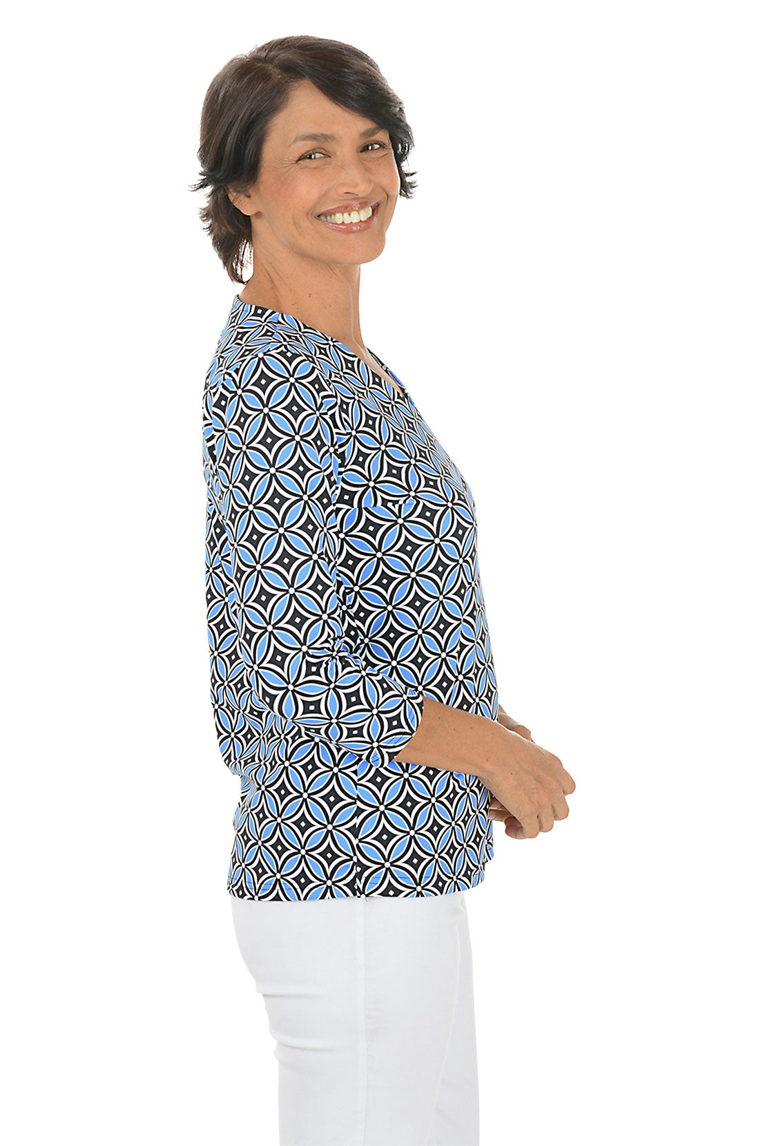 Woman wearing a geometric patterned blue and black blouse with white pants on a white background. Side shot.