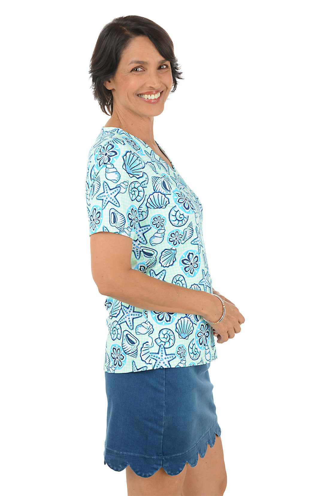 Woman wearing a blue patterned blouse with a shell design and a scallop denim skort on a white background. Side shot.