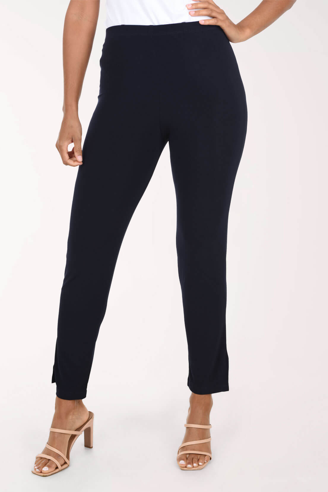 A model wearing slim-fit navy ankle-length pants with side slits, paired with a white top and beige strappy heeled sandals, shown against a plain white studio background.