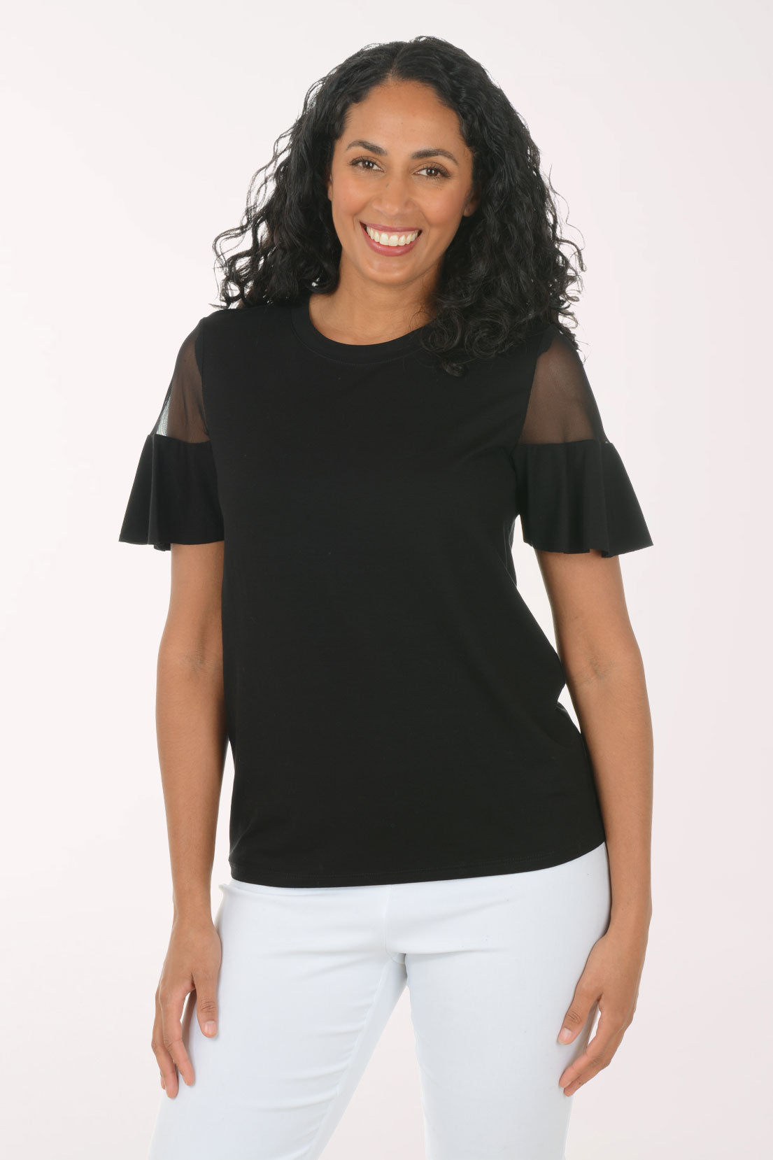 Woman wearing a black top with sheer sleeves and white pants on a light pink background