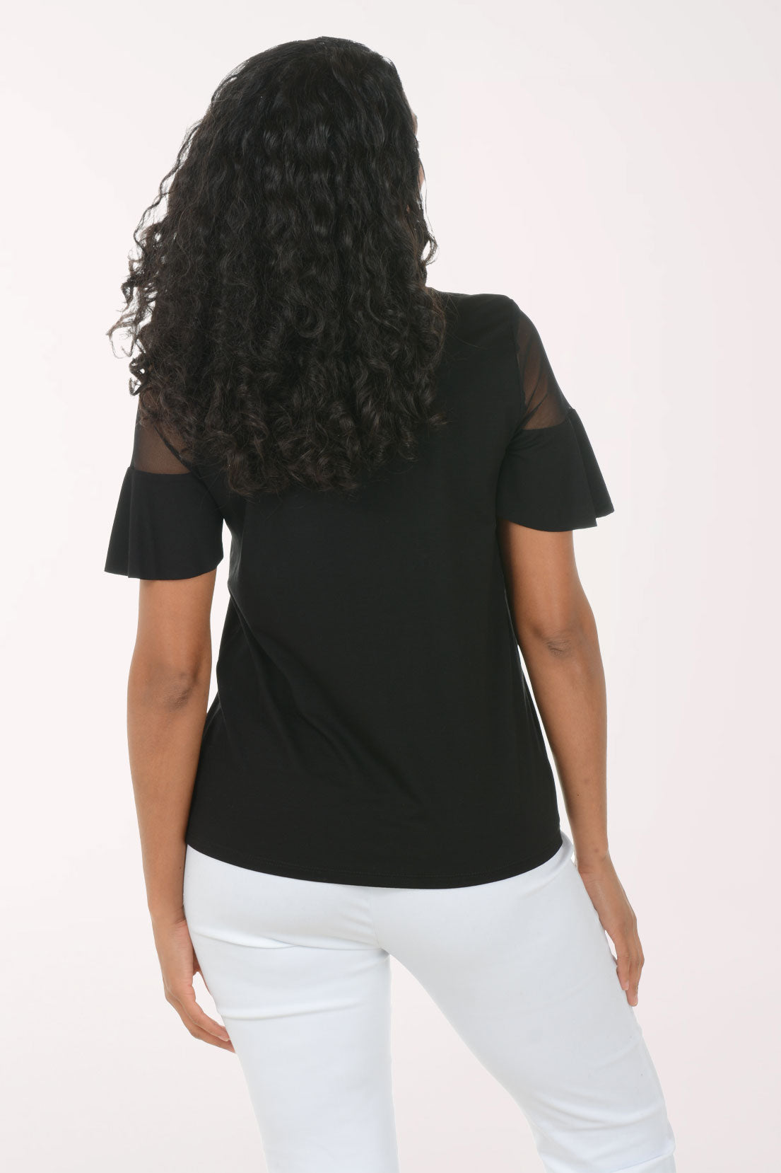 Woman wearing a black top with sheer sleeves and white pants on a light pink background