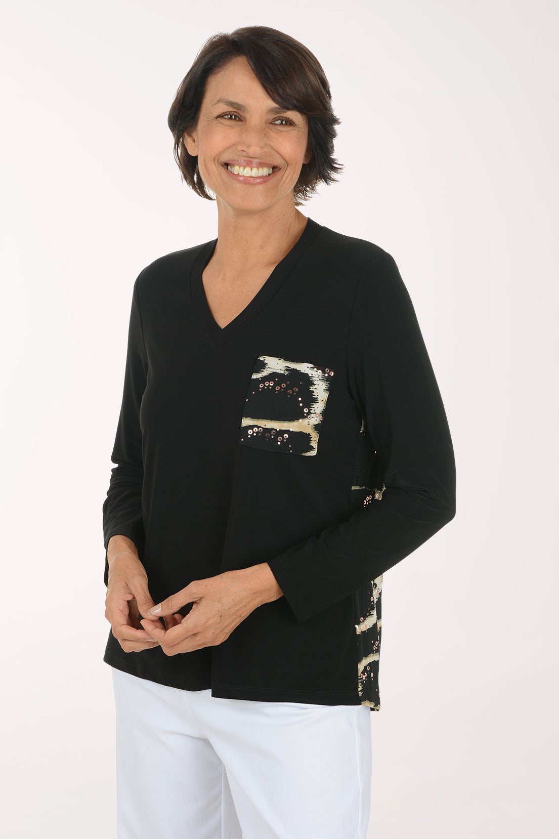 A smiling woman wearing a black long-sleeve V-neck sweater with a sequined abstract chest design in beige, white, and black, paired with white pants, against a plain white studio background.