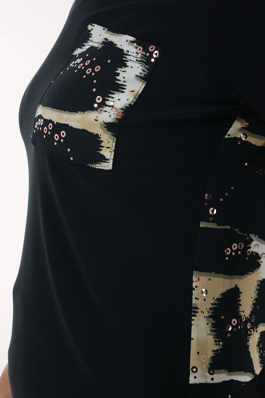 A smiling woman wearing a black long-sleeve V-neck sweater with a sequined abstract chest design in beige, white, and black, paired with white pants, against a plain white studio background.