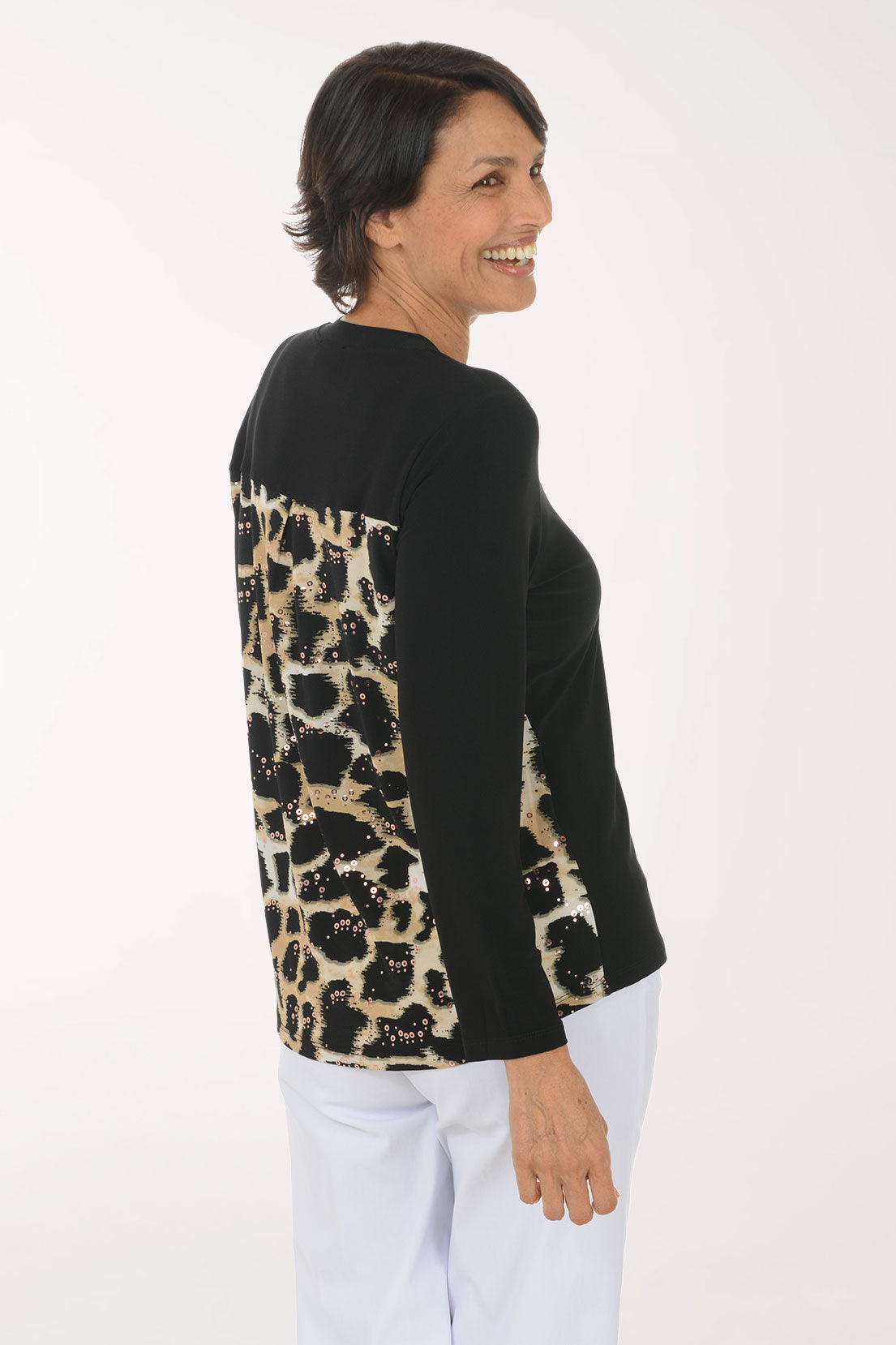 A smiling woman wearing a black long-sleeve V-neck sweater with a sequined abstract chest design in beige, white, and black, paired with white pants, against a plain white studio background.