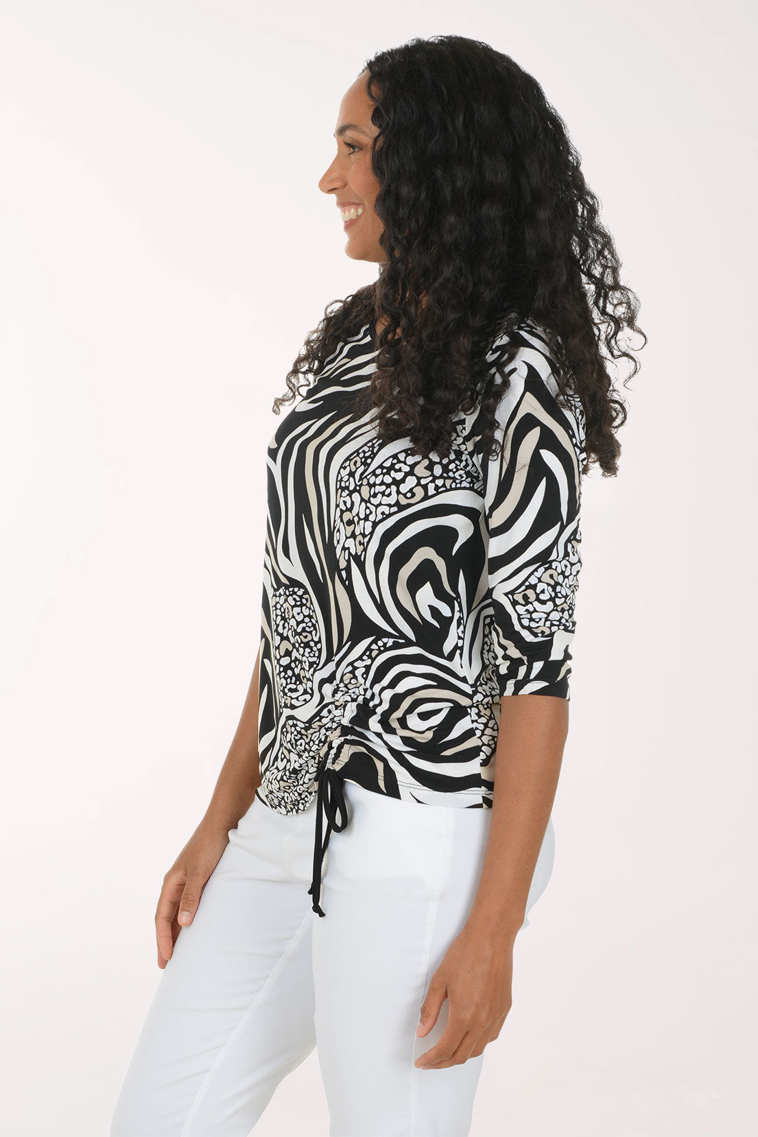A woman modeling a black and white zebra print top with three-quarter sleeves and a side tie, paired with white pants, against a plain light background.