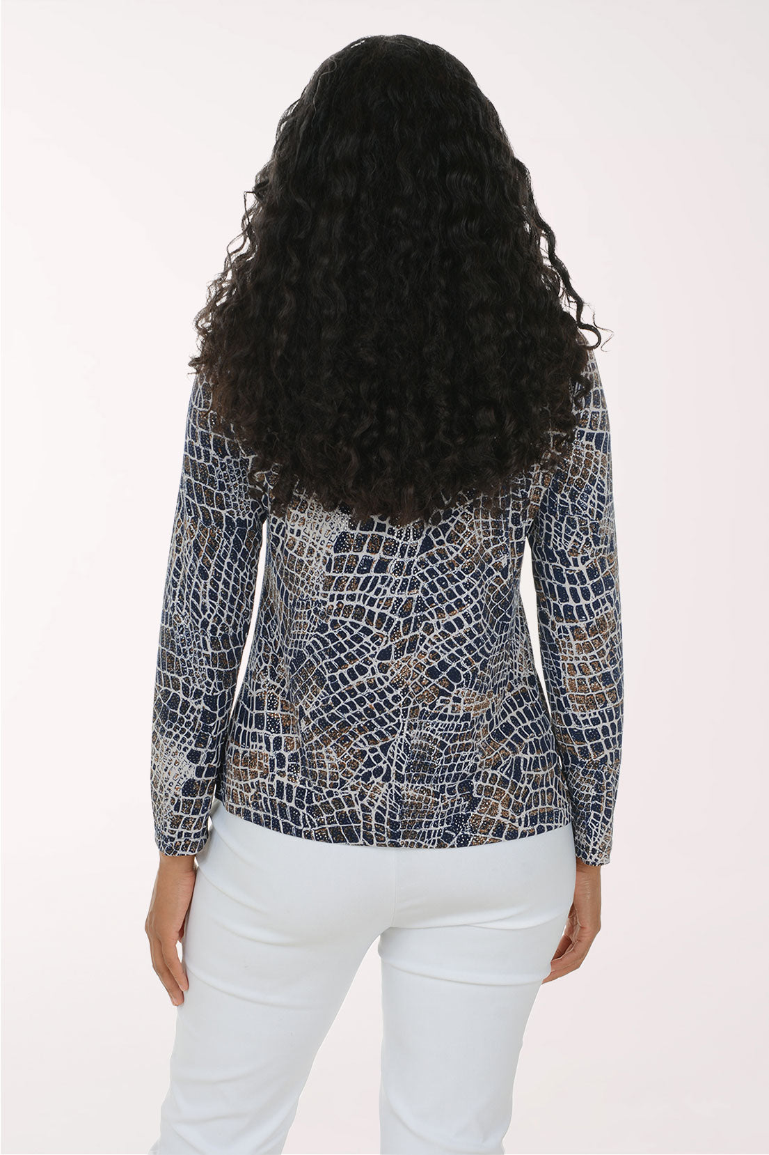 A woman modeling a long-sleeve V-neck top with a blue, white, and beige reptile or snakeskin pattern, paired with white pants, against a plain white studio background.