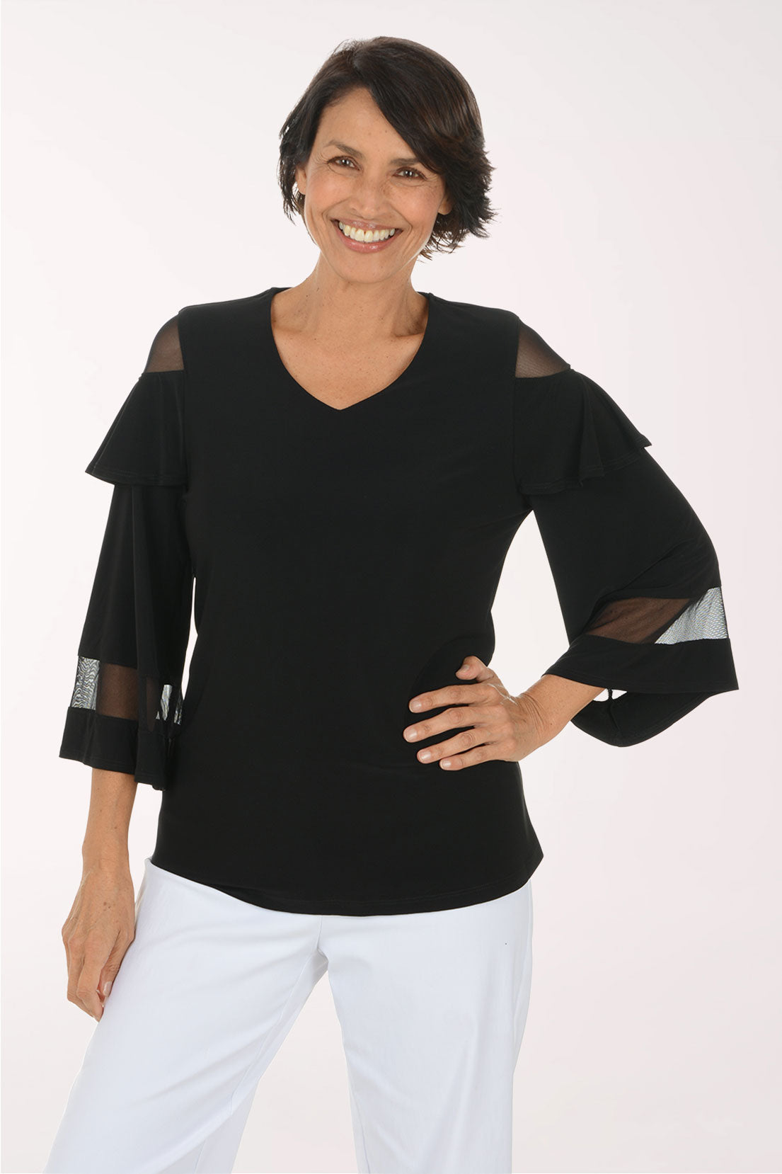 A smiling woman wearing a black V-neck top with sheer mesh shoulder and elbow panels, paired with white pants, photographed against a plain light background.